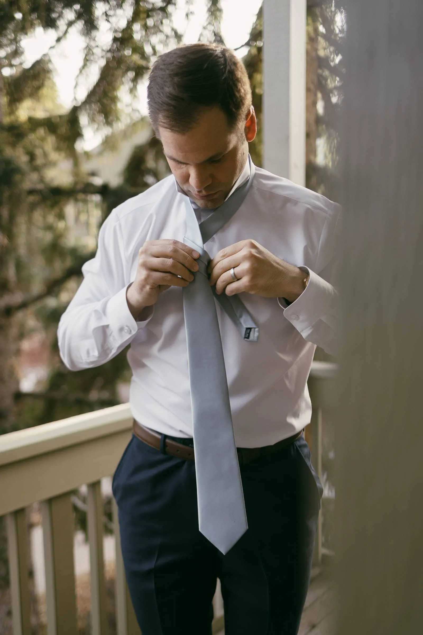  A man in a white shirt and dark pants stands on a porch, looking down and tying a light gray necktie, with trees visible in the background. 