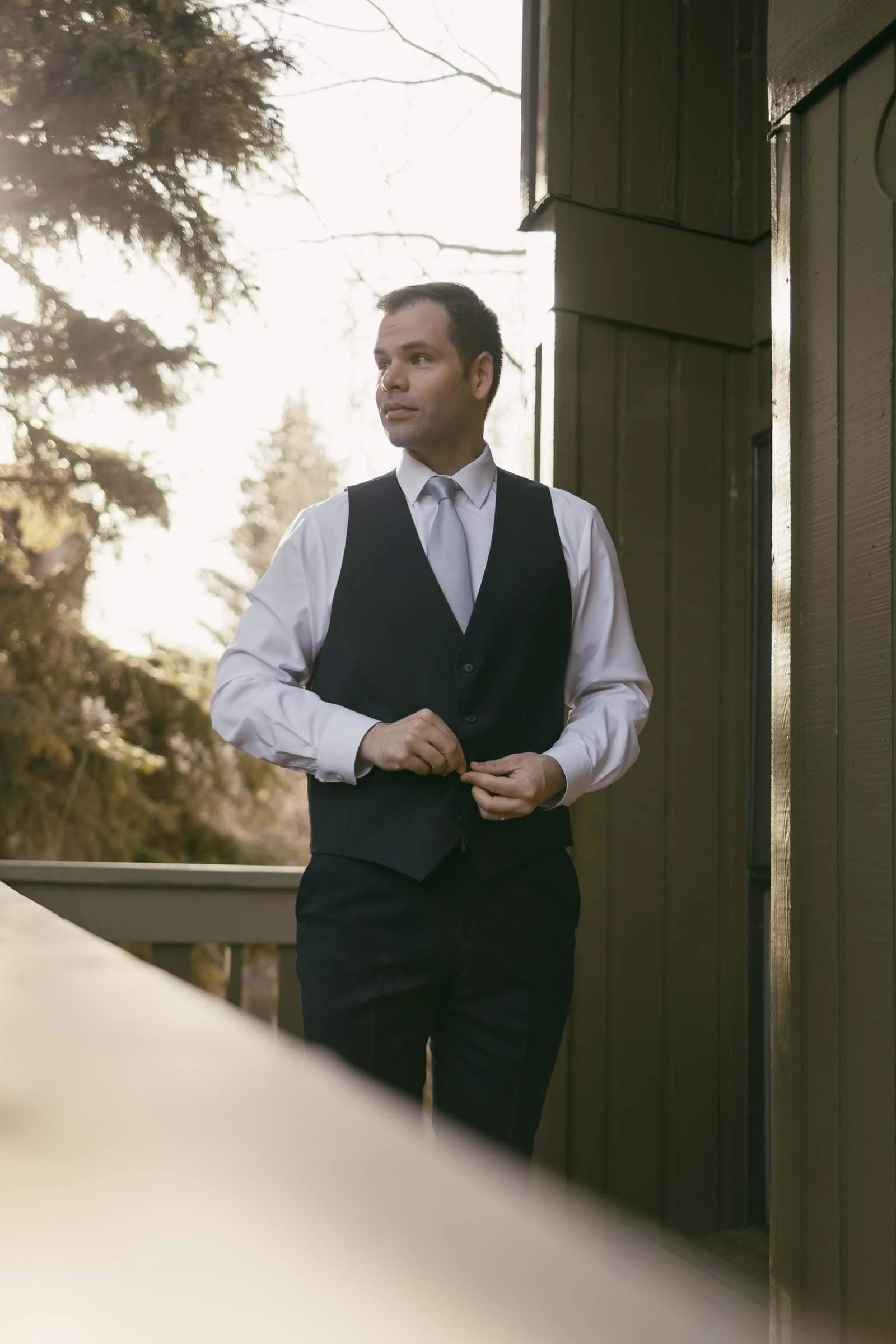  A man in a white shirt and dark vest stands on a balcony, buttoning his vest. He looks to the side thoughtfully, with trees and soft sunlight in the background. 