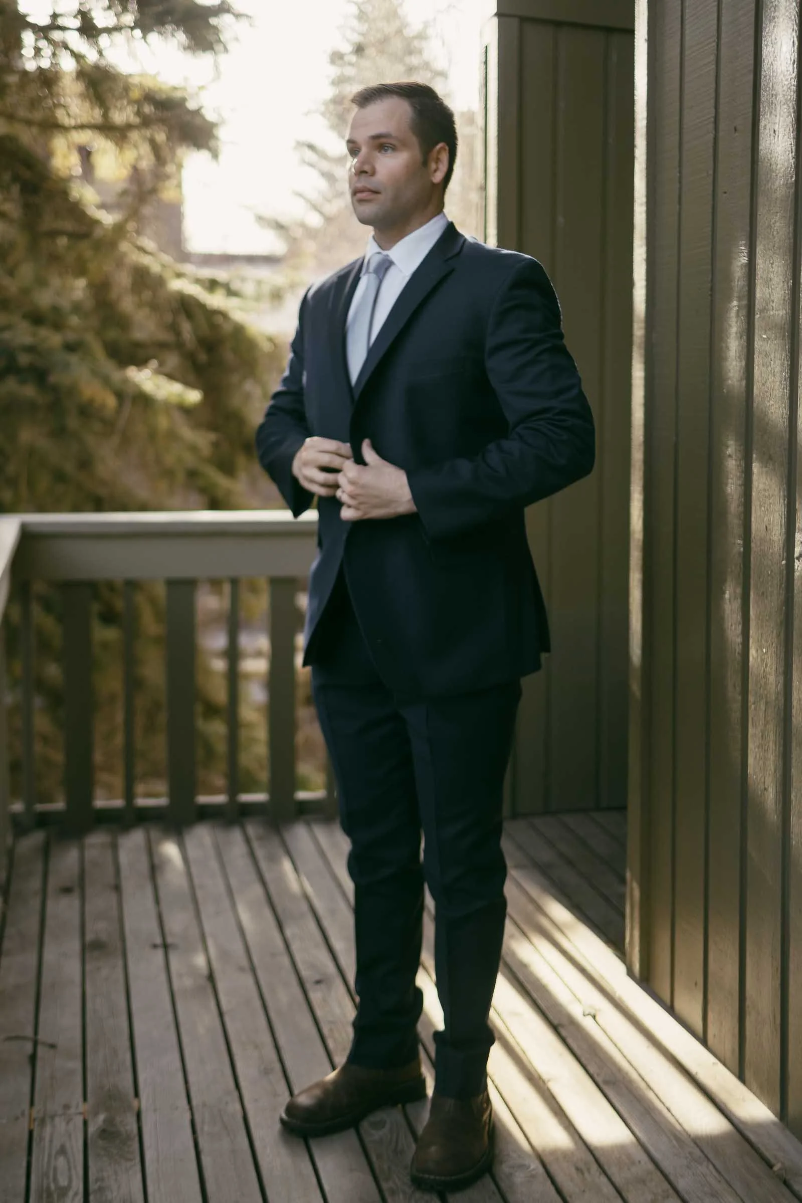  A man in a navy suit and tie stands on a wooden deck, buttoning his jacket. He is outdoors, with sunlight filtering through trees in the background. 