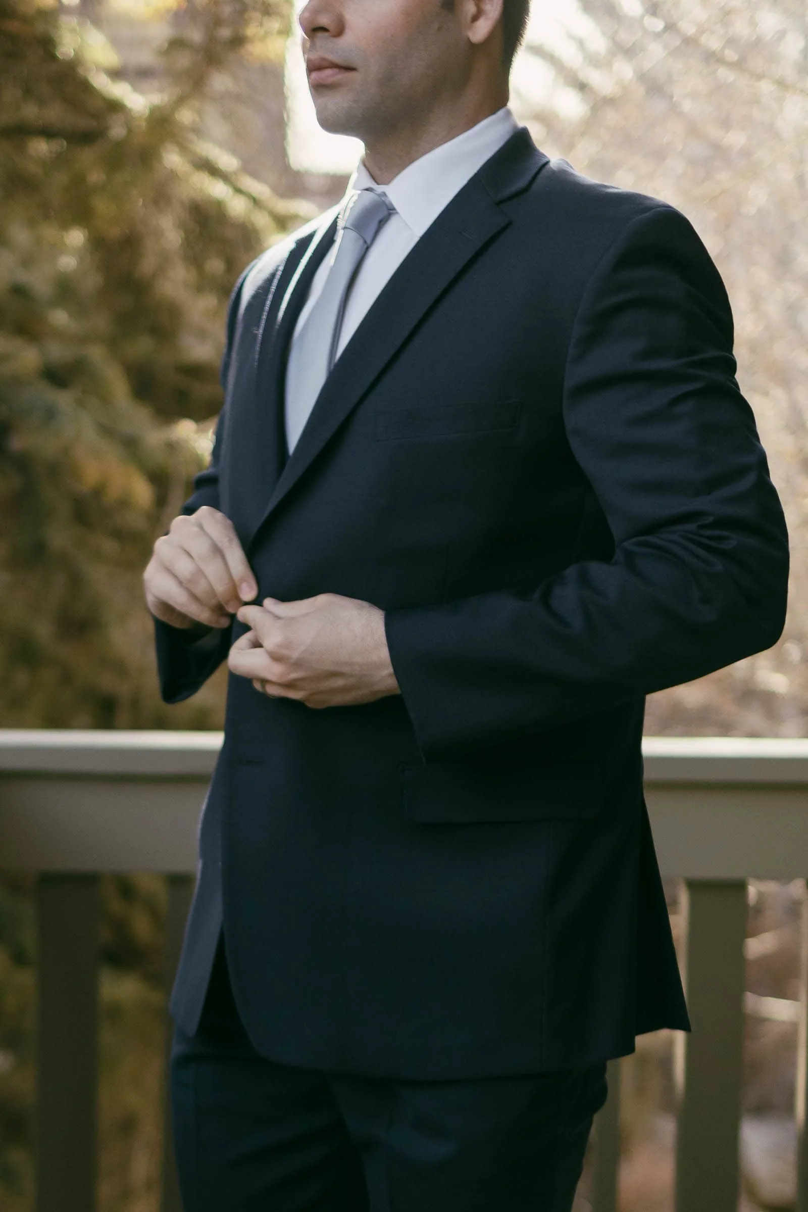  A man in a dark suit and tie stands outdoors, buttoning his jacket. His face is partially out of frame, and the background shows blurred trees and a wooden railing in soft sunlight. 