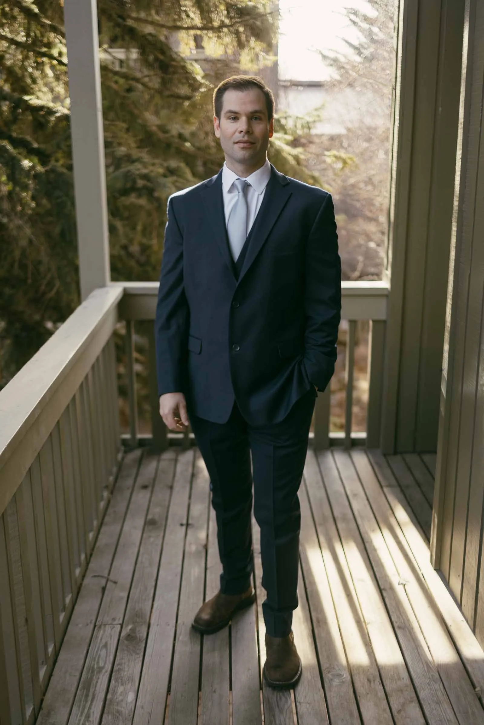  A man in a navy suit, white shirt, and tie stands on a wooden balcony with one hand in his pocket. Sunlight filters through trees in the background. 