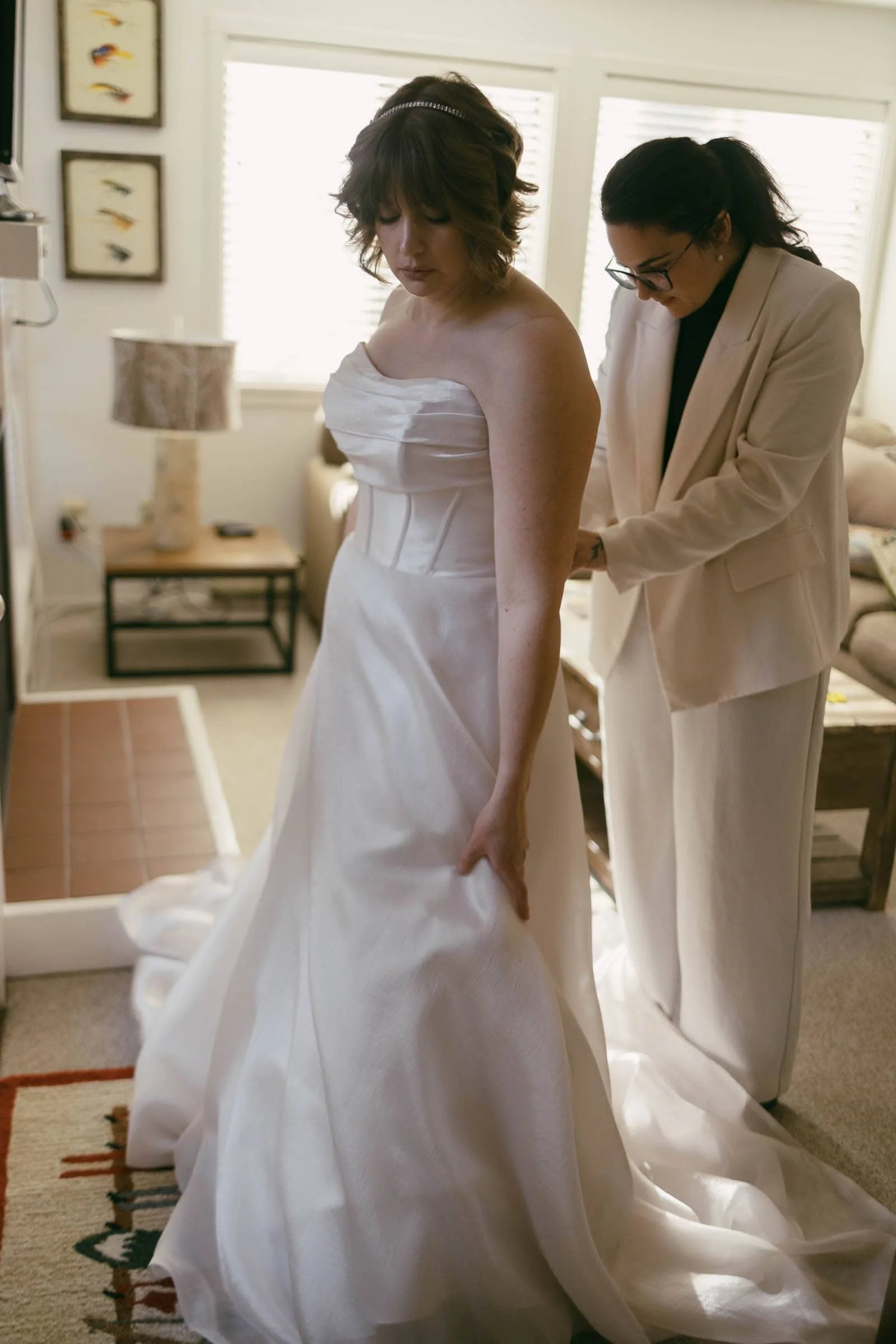  A bride in a strapless white gown stands indoors while another woman in a light-colored suit helps adjust her dress. The room is softly lit with natural light from windows in the background. 