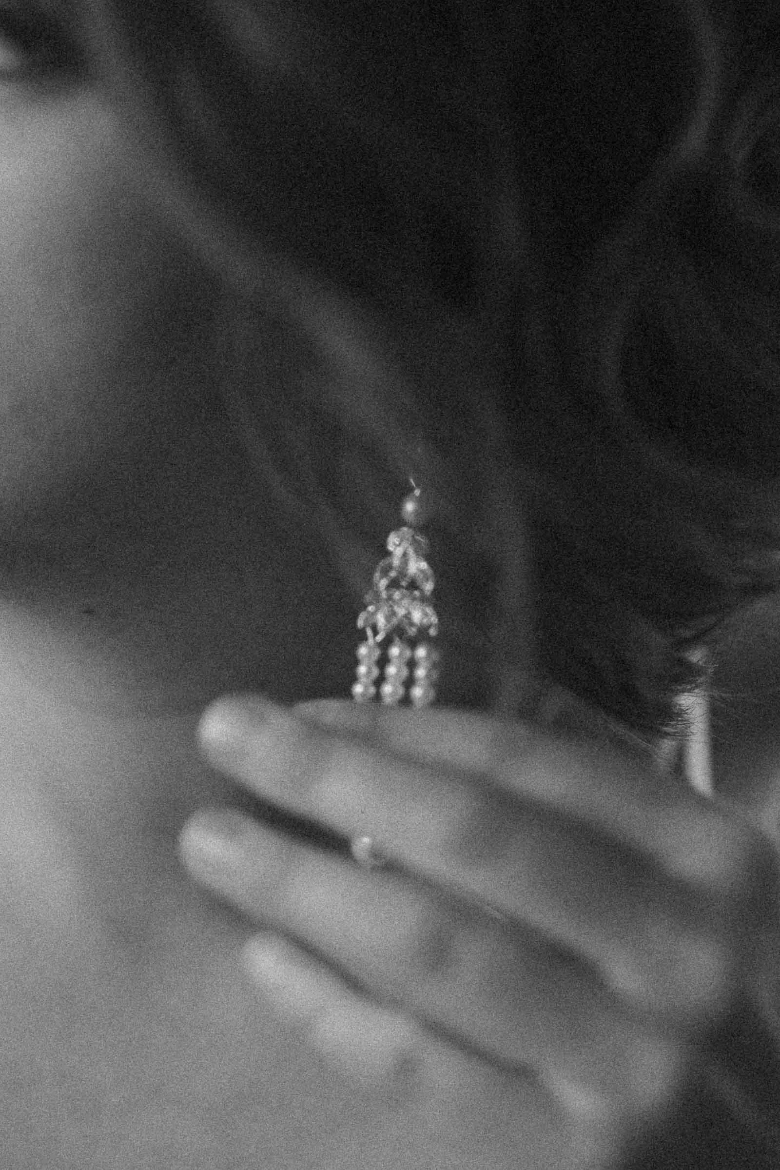  Black and white close-up of a person holding a long, dangling earring with pearls and decorative elements near their ear. The image is softly focused, highlighting the jewelry and the person's hand. 
