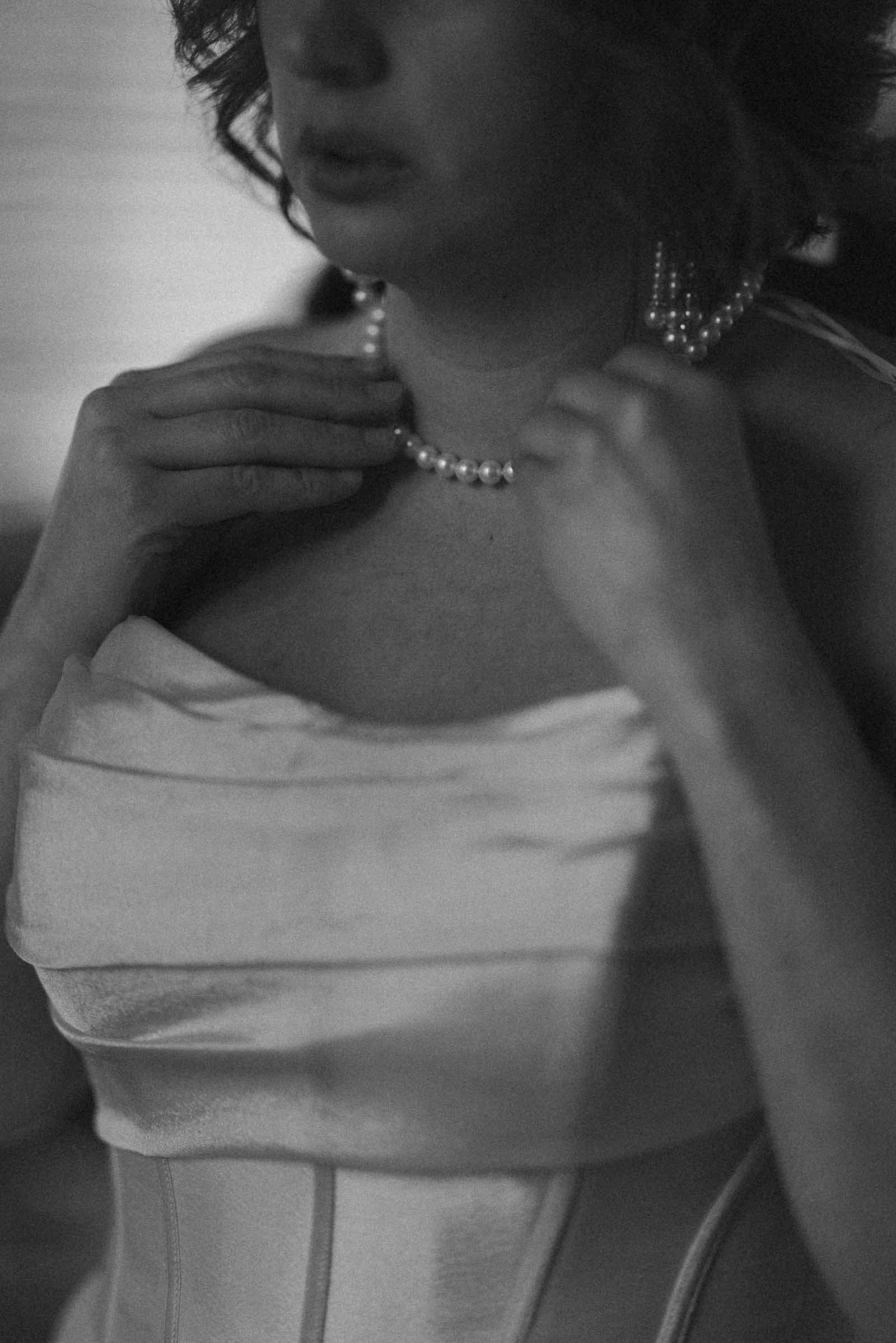  A woman in a strapless dress adjusts a pearl necklace around her neck. The photo is in black and white and slightly out of focus, creating a soft, intimate atmosphere. 