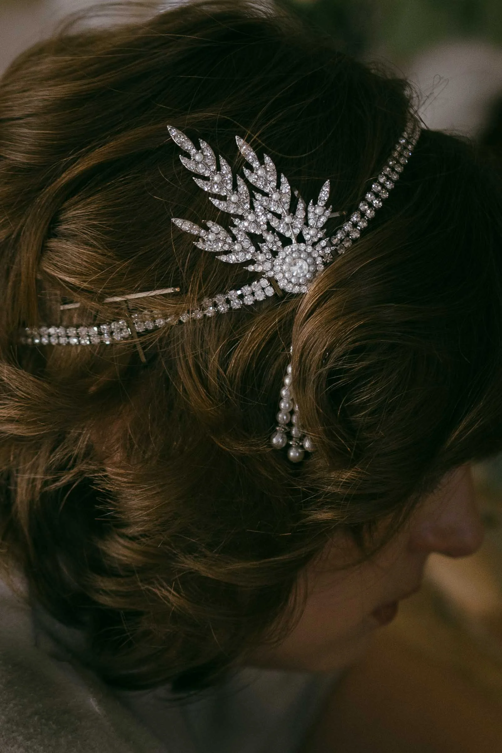  Close-up of a person with brown wavy hair wearing an ornate, silver hair accessory adorned with sparkling gems and pearls in a leaf-like design. The face is partially out of focus and only partly visible. 