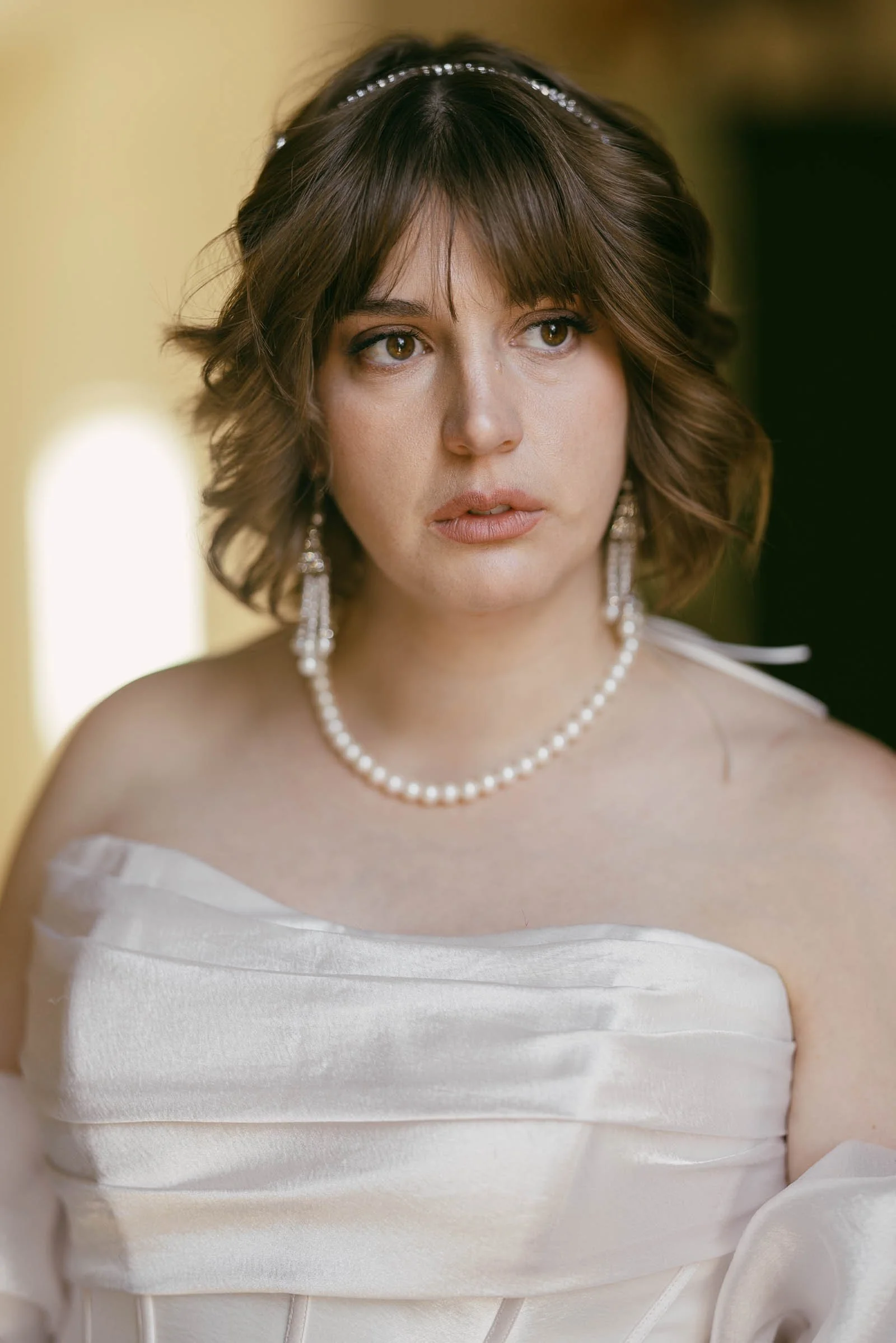  A woman wearing a white strapless dress with a pearl necklace and earrings, and a headband in her wavy brown hair, looks thoughtfully to the side. The background is softly blurred. 