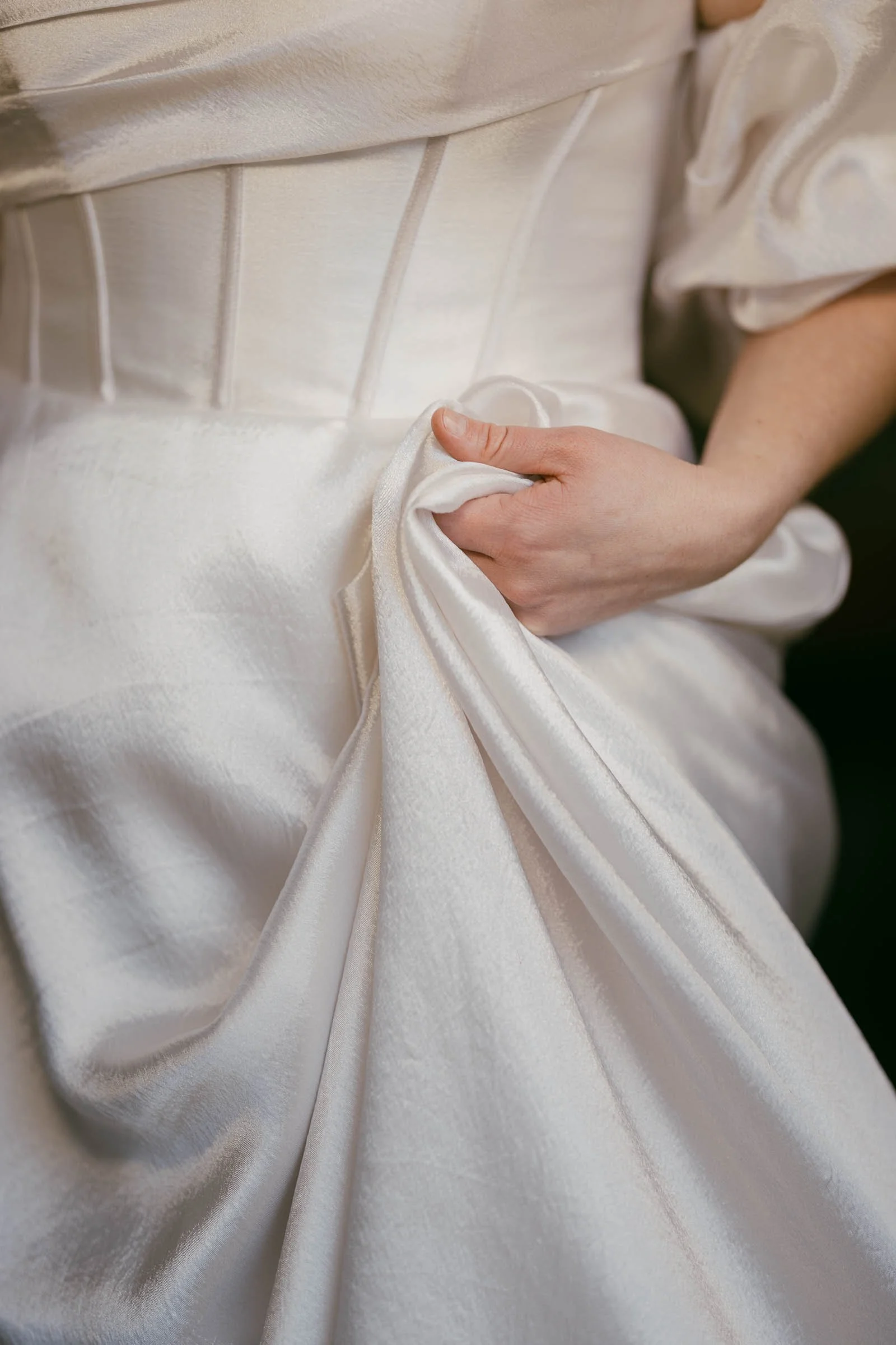  Close-up of a person in a white satin dress, holding a handful of the fabric in one hand. The dress features a structured bodice and puffed sleeves. The image focuses on the texture and detail of the clothing. 