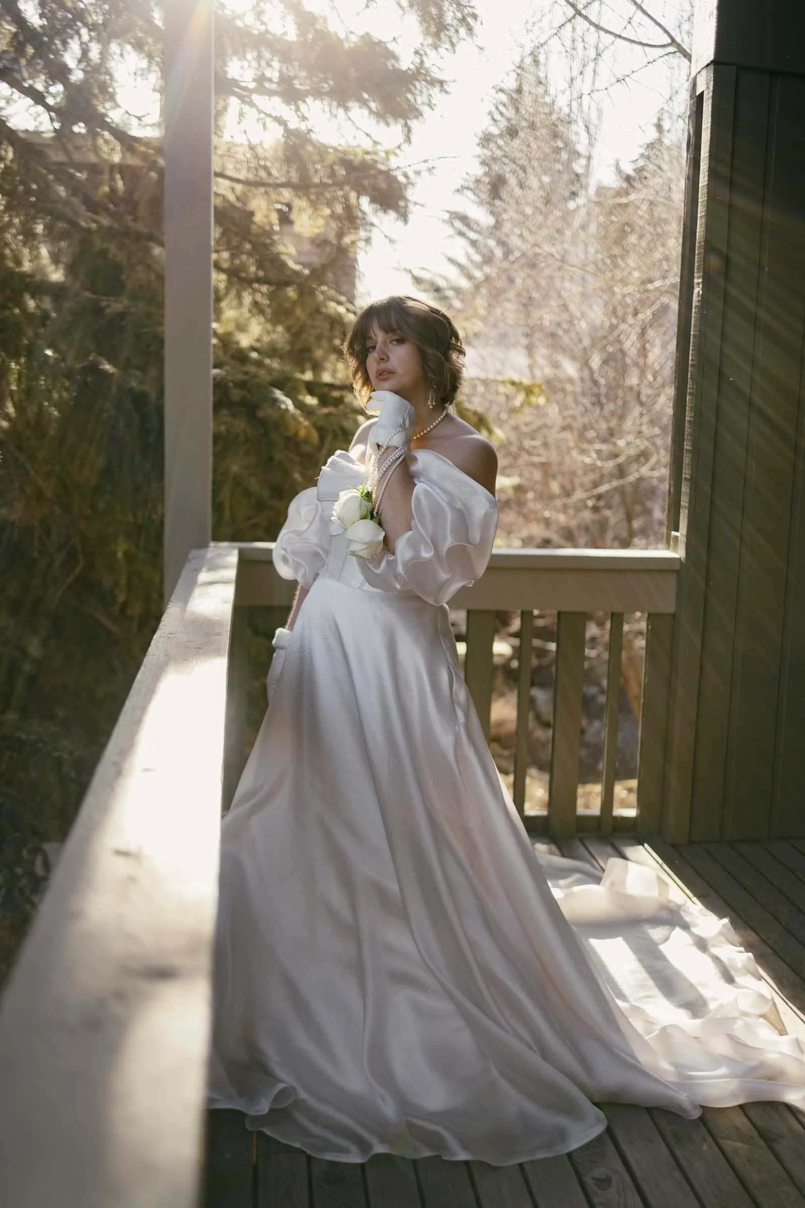  A woman in a flowing white off-the-shoulder gown stands on a wooden balcony glowing in sunlight, holding a white rose while gazing thoughtfully into the distance. Tall trees and warm filtered light create a soft natural backdrop behind her. 