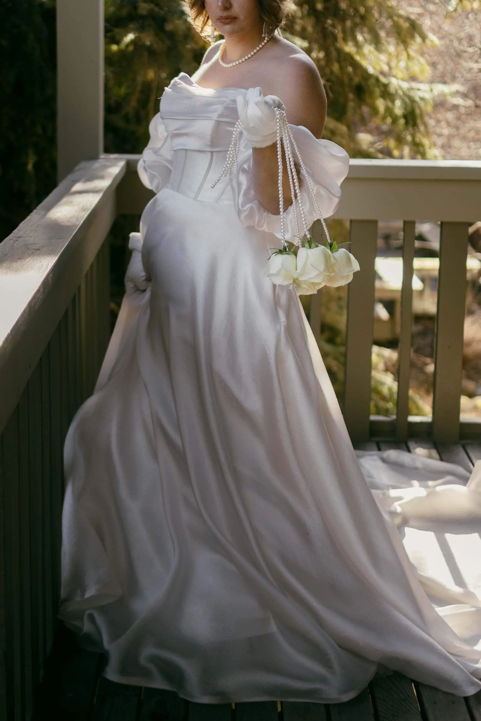  A person wearing an off-the-shoulder white satin gown and pearl necklace stands on a wooden porch, holding a string of white roses. The background features greenery and sunlight. The person's face is not visible. 