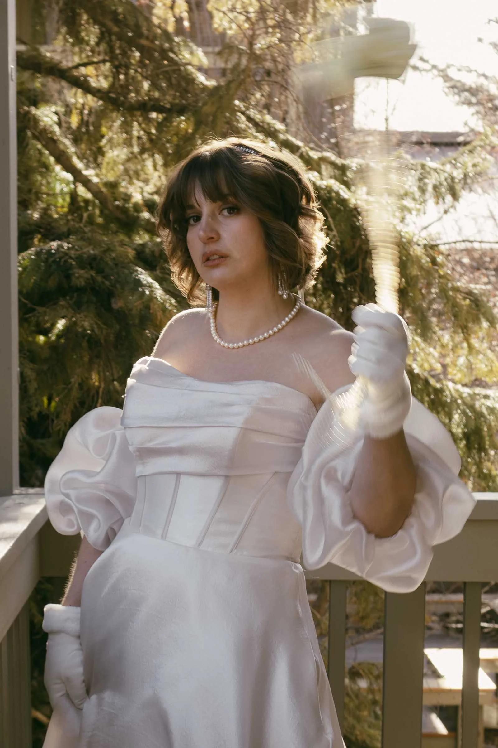  A person in a white, off-the-shoulder gown with puffed sleeves, gloves, and a pearl necklace stands on a porch, holding a blurred handheld fan. Pine trees and sunlight are visible in the background. 
