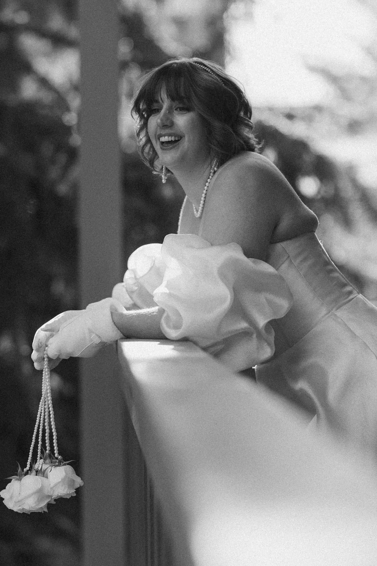  A woman in a formal gown and gloves leans on a railing, smiling. She holds a string with white roses and wears pearl jewelry. The image is in black and white with soft, natural lighting. 