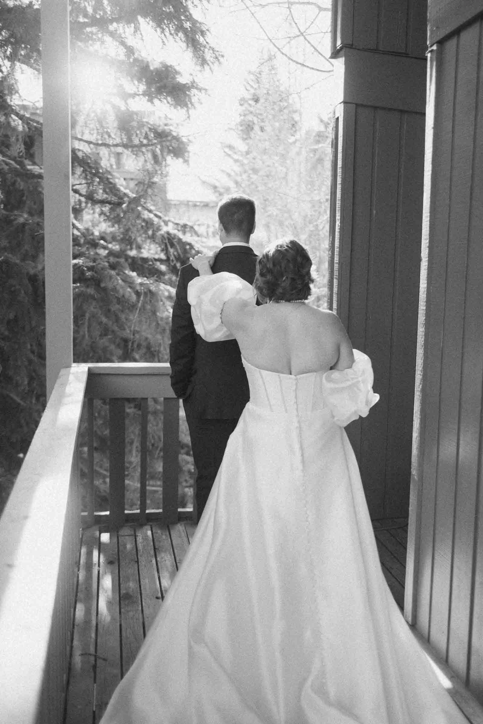  A bride in a white dress approaches a groom standing on a wooden balcony, ready for a first look moment; sunlight filters through trees in the background. 
