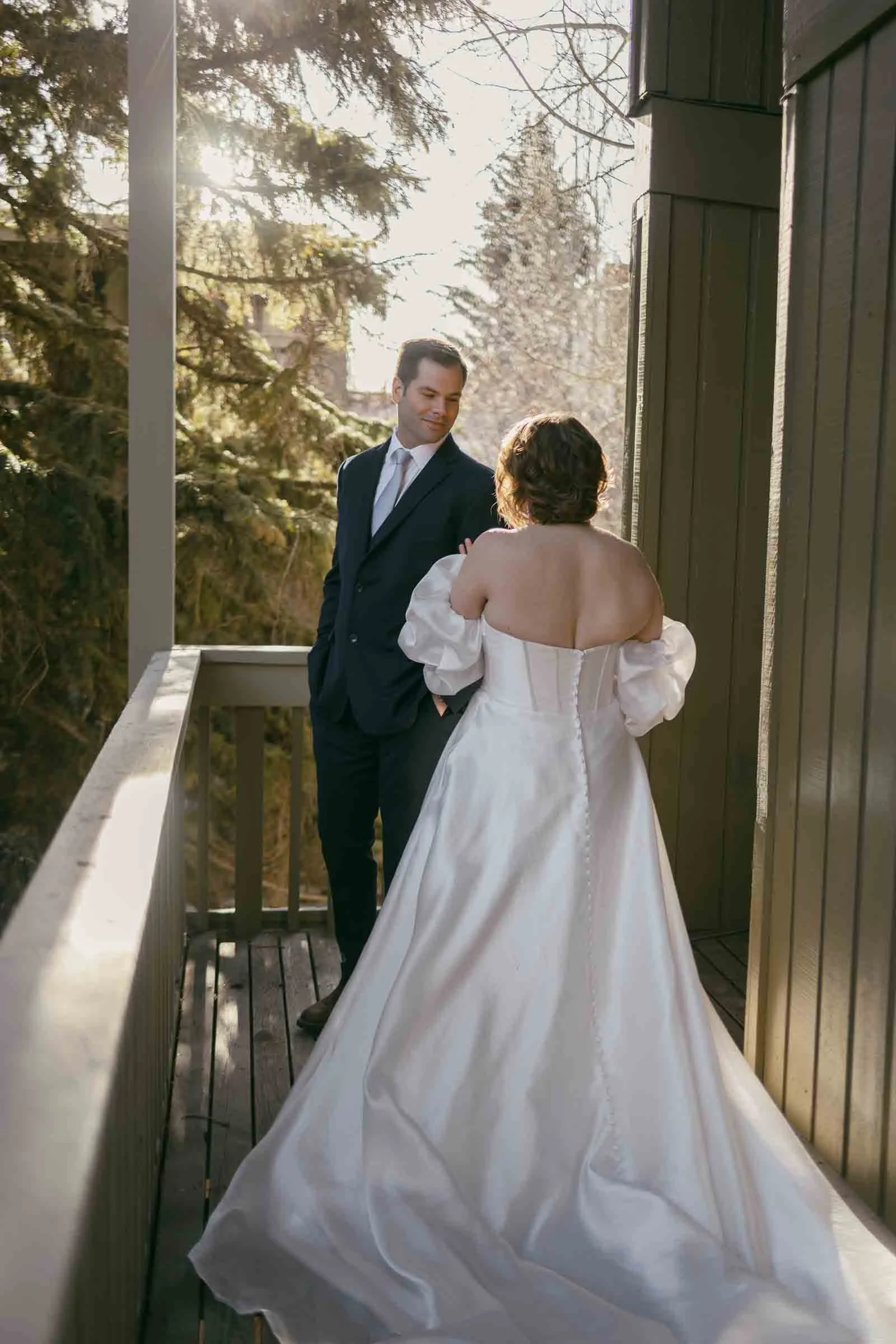  A bride wearing a white dress with puffed sleeves stands on a wooden balcony facing a man dressed in a suit. Golden sunlight filters through the tall trees behind them as the two share a quiet outdoor moment together. 