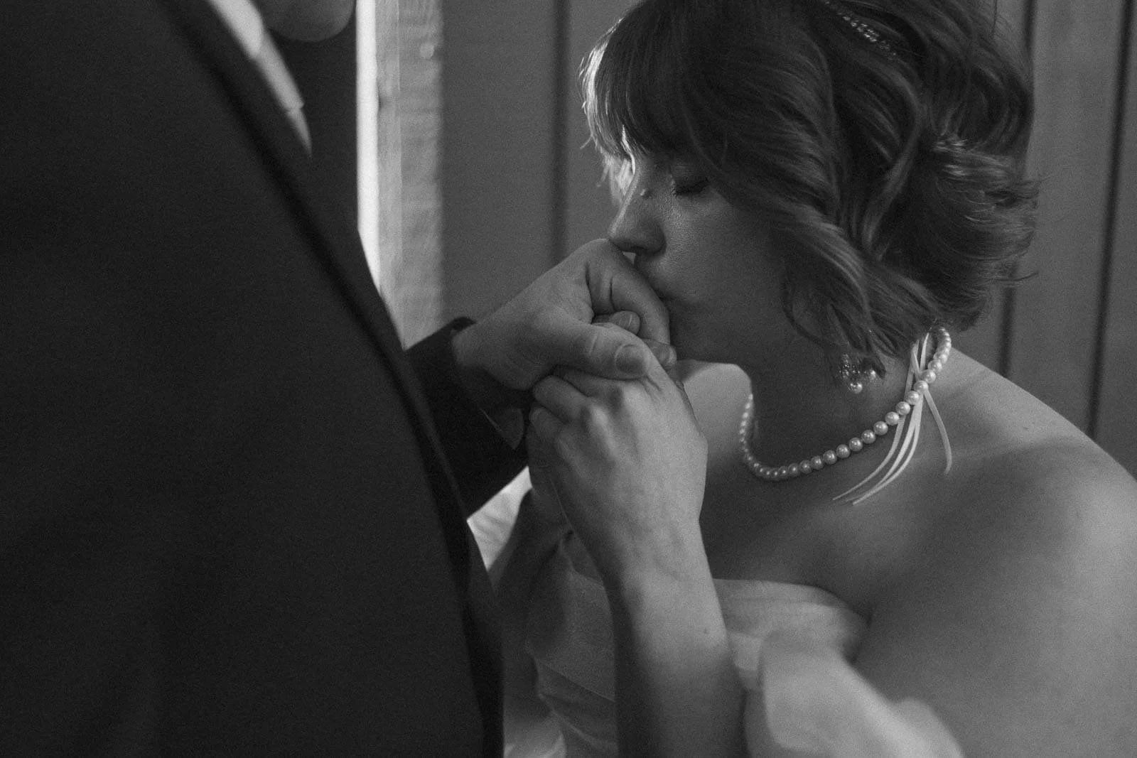  A bride in a strapless dress and pearl necklace gently kisses the hand of a person in a suit, capturing an intimate and emotional moment between them. 