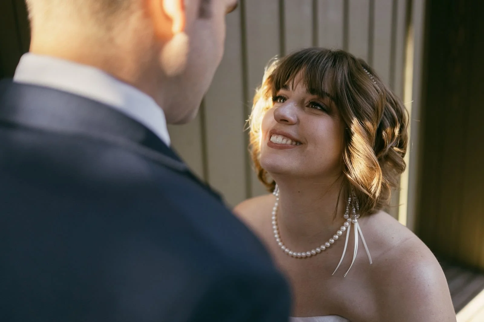  A woman in a white dress and pearl necklace smiles warmly up at a man in a suit, standing close together in soft sunlight by a wooden wall. 