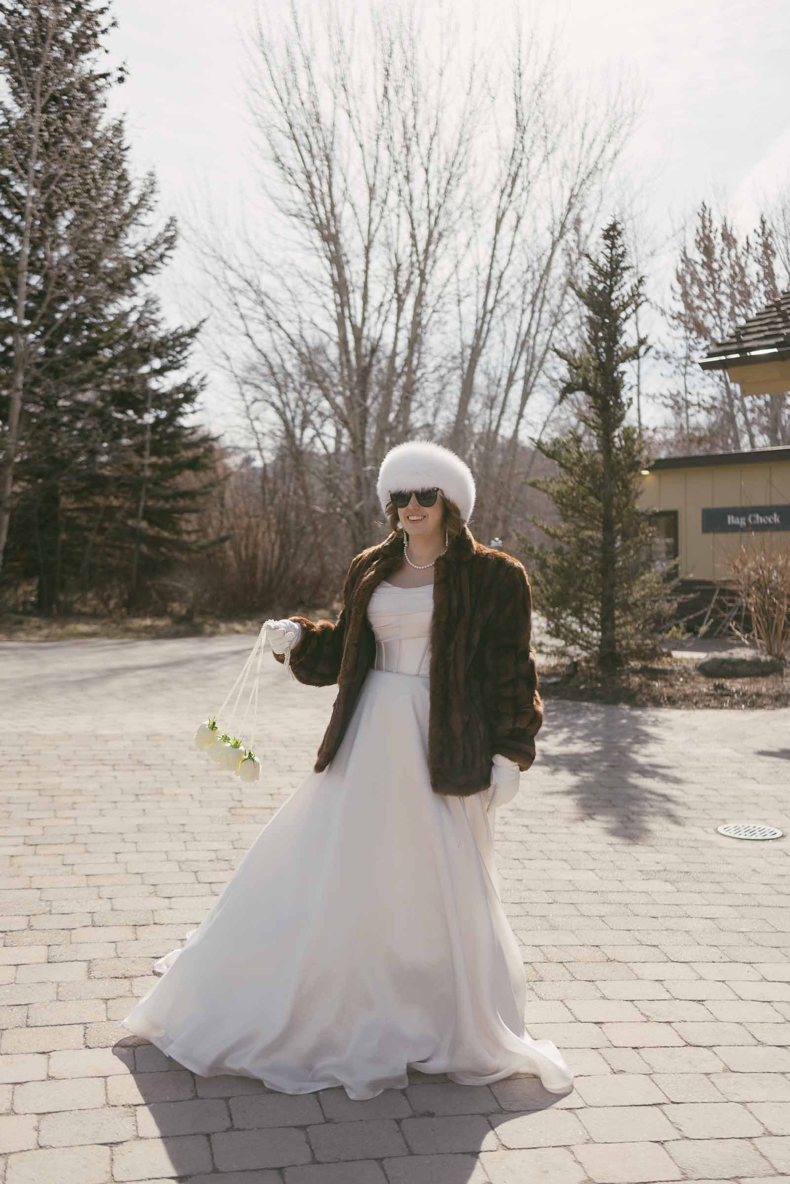  A bride in a white gown, fur coat, white hat, and sunglasses stands outdoors on a paved walkway, holding a small bouquet, with trees and a building in the background. 