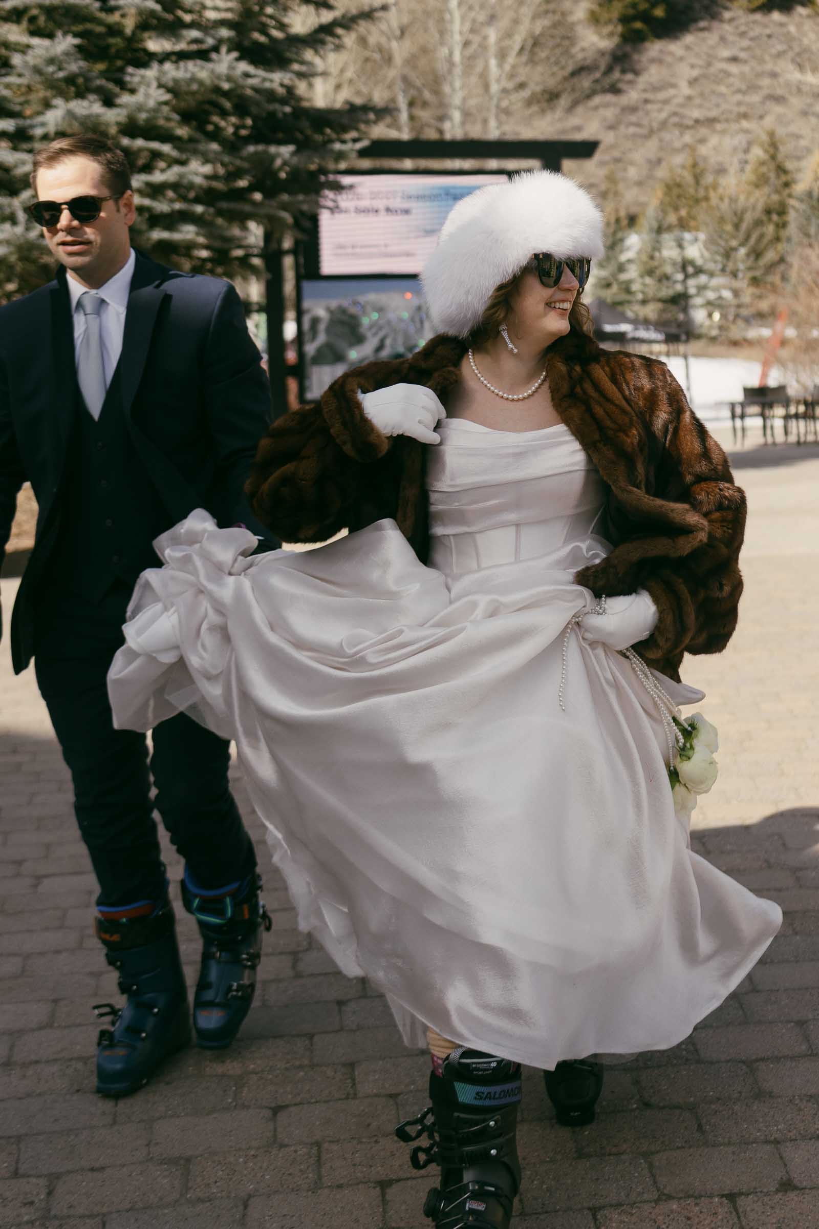  A bride in a white dress, fur coat, and white furry hat walks beside a man in a suit. Both wear ski boots and sunglasses, outdoors on a sunny day with trees and mountains in the background. 