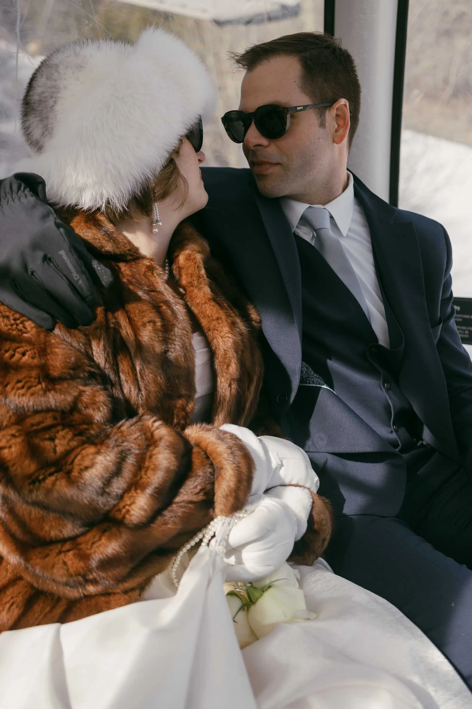  A couple dressed in elegant winter attire sits closely together. The woman wears a fur coat, white gloves, and a white fur hat, while the man wears a dark suit and sunglasses. They look at each other lovingly inside a gondola. 
