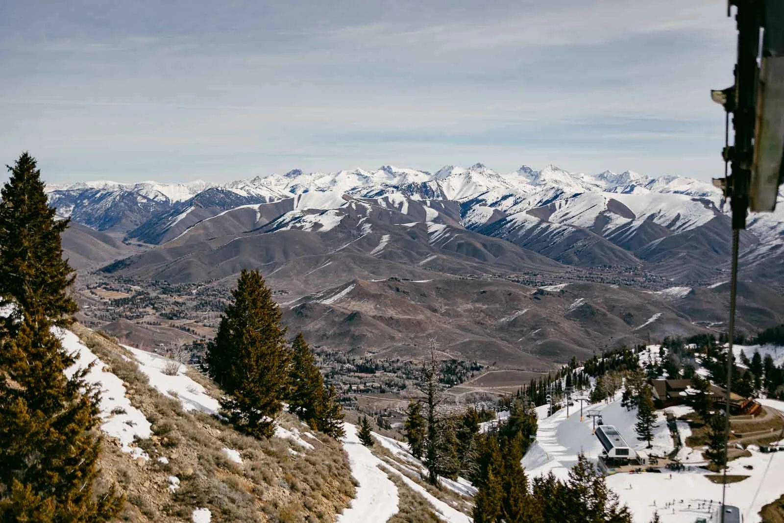  A scenic view of snow-capped mountains and rolling hills, with pine trees in the foreground and patches of snow covering the ground under a cloudy sky. A ski lift and small buildings are visible in the lower right corner. 