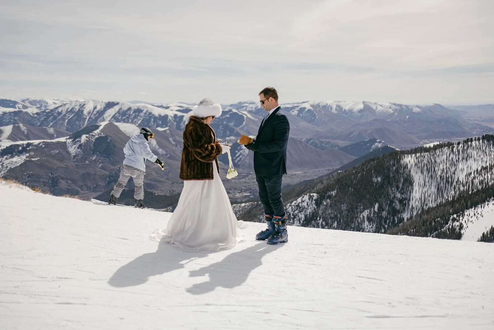 A bride and groom stand on a snowy mountain while exchanging vows. The bride wears a white dress with a fur coat, and the groom wears a suit paired with ski boots. A snowboarder glides past them as snow-covered mountains fill the background.