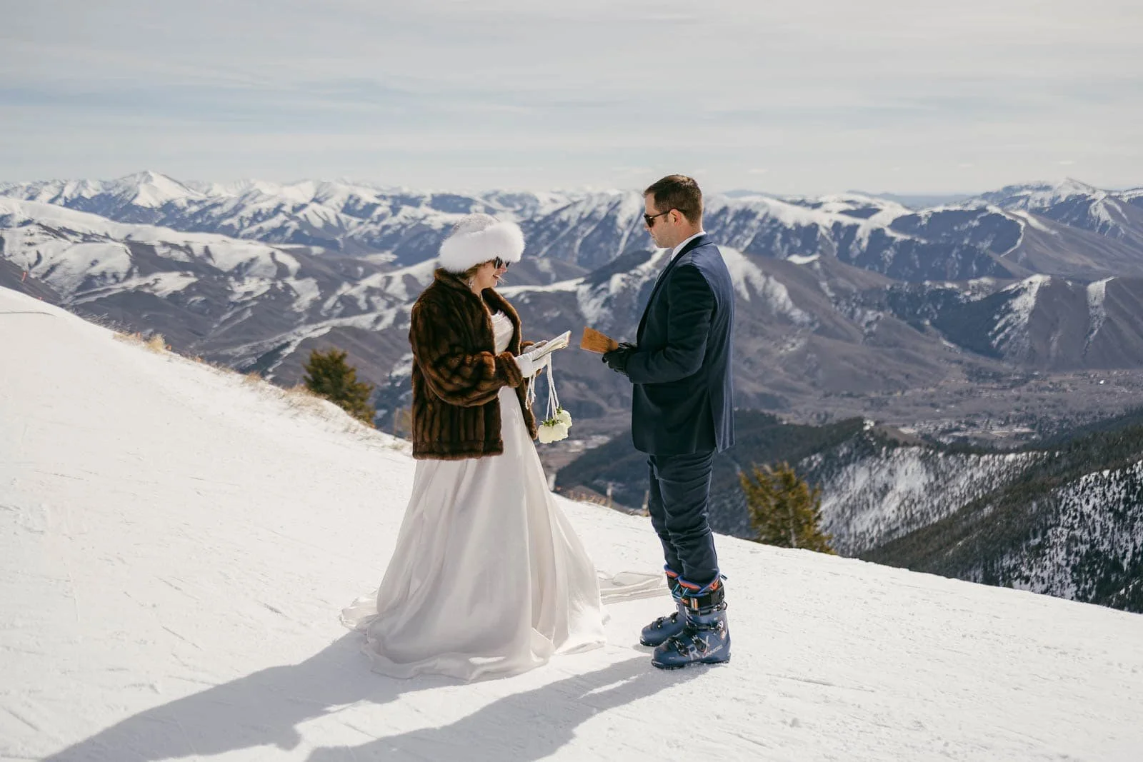  A bride and groom exchange vows on a snowy mountaintop with snow-covered mountains in the background. The bride wears a fur hat and coat, while the groom wears a suit and ski boots. 