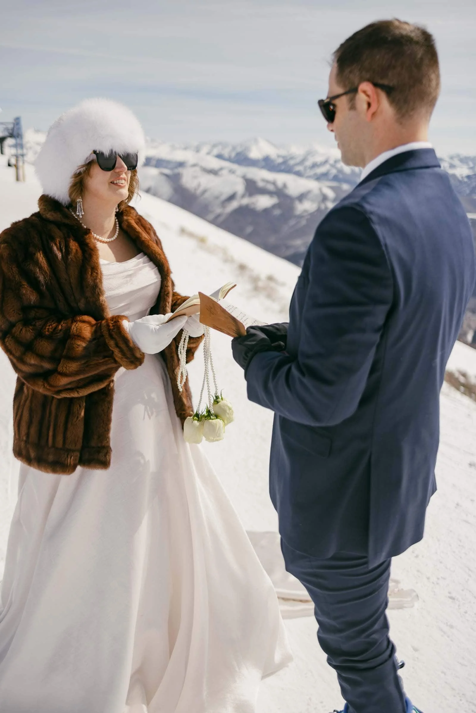  A bride and groom stand together outdoors in the snow with rugged mountain peaks rising in the background. The bride wears a white dress, fur coat, and fluffy winter hat, while the groom wears a dark suit. Both wear sunglasses as they read their vow