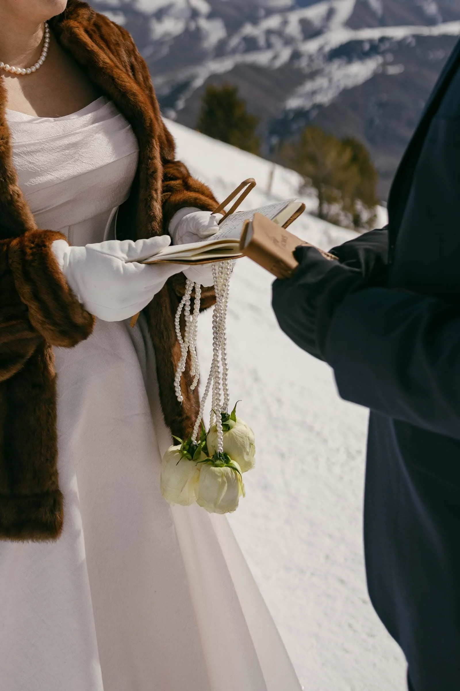  A bride in a white dress, fur coat, and gloves holds a book and white flowers on cords while standing with a suited partner on a snowy mountain. Only their torsos and hands are visible. 