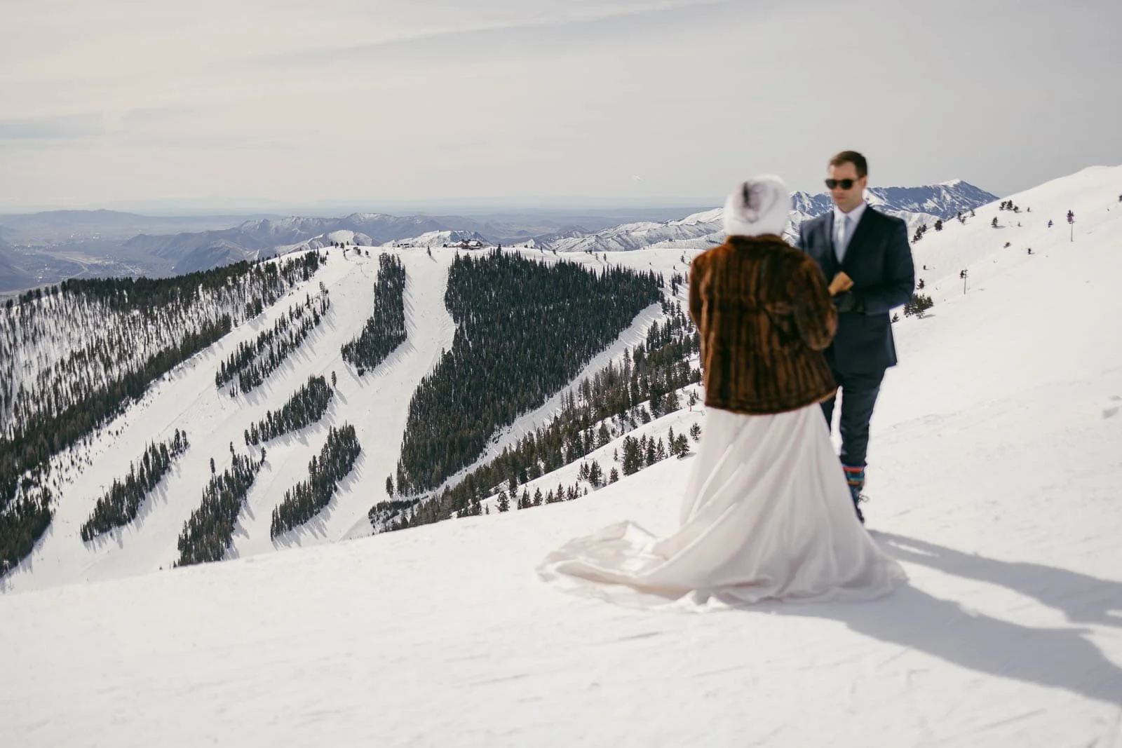  A bride and groom stand together on a snowy mountain slope, with ski trails and snow-covered trees visible in the background under a cloudy sky. 