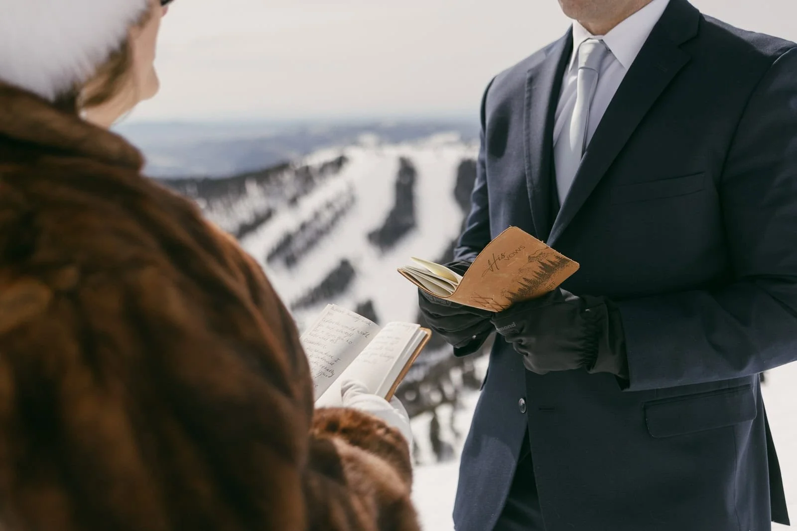  Two people dressed in formal winter clothing stand outside on a snowy mountain holding vow books while facing one another, exchanging a meaningful moment together. 