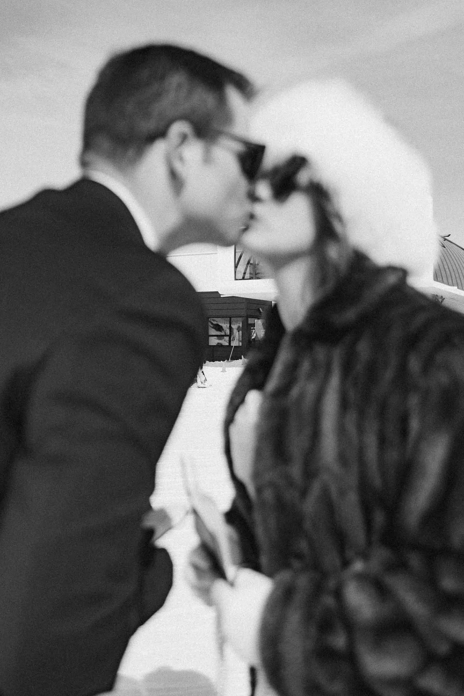  A black-and-white photo shows a man and woman outdoors in winter attire, sharing a kiss. The background is snowy, and both wear sunglasses; the woman has a fur hat and coat, while the man is in a suit. The image is slightly out of focus. 