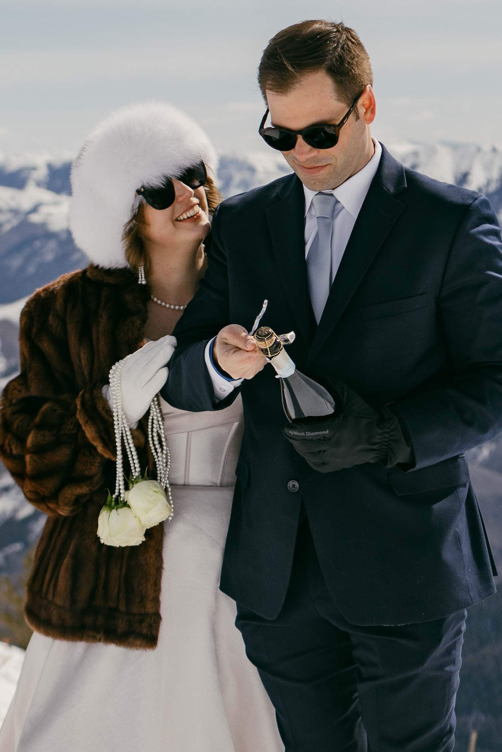 A bride in a white dress, fur coat, and white hat stands beside a groom in a dark suit and sunglasses. He opens a bottle of champagne in a snowy mountain setting. The bride holds pearls and white flowers, smiling at him. 