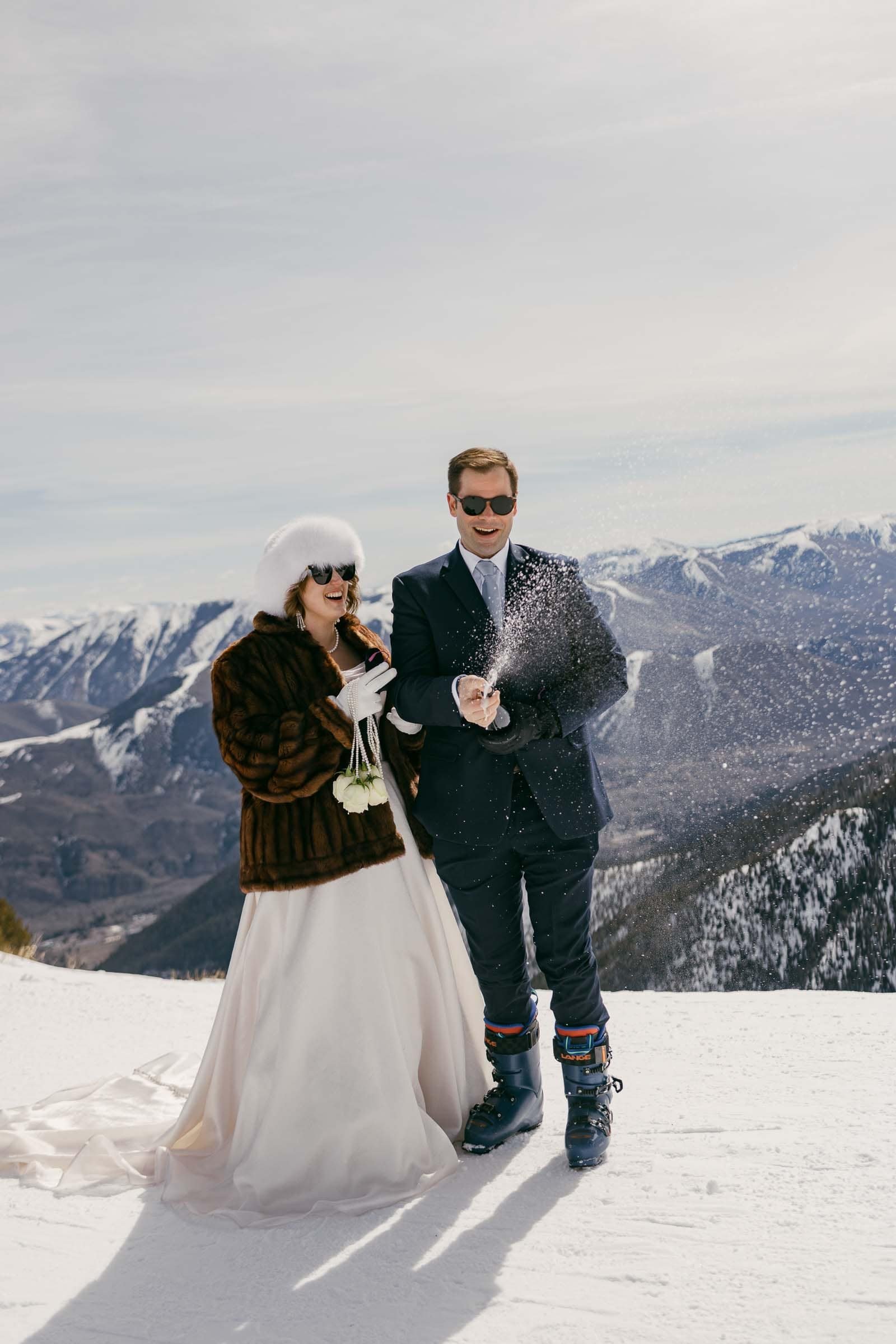  A bride in a white dress and fur coat and a groom in a suit and ski boots celebrate on a snowy mountain, with the groom popping a bottle of champagne as snow-capped peaks stretch out in the background. 