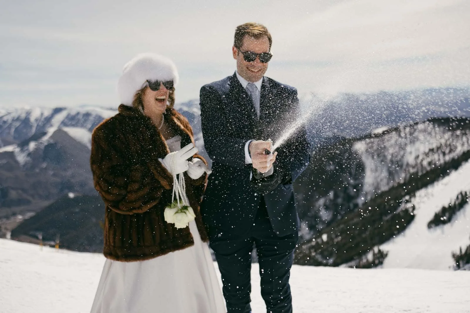 A bride and groom bundled in winter attire stand on a snowy mountain, laughing as the groom opens a bottle of champagne, sending bubbles spraying into the cold mountain air. Snowy peaks and a bright blue sky stretch across the background.