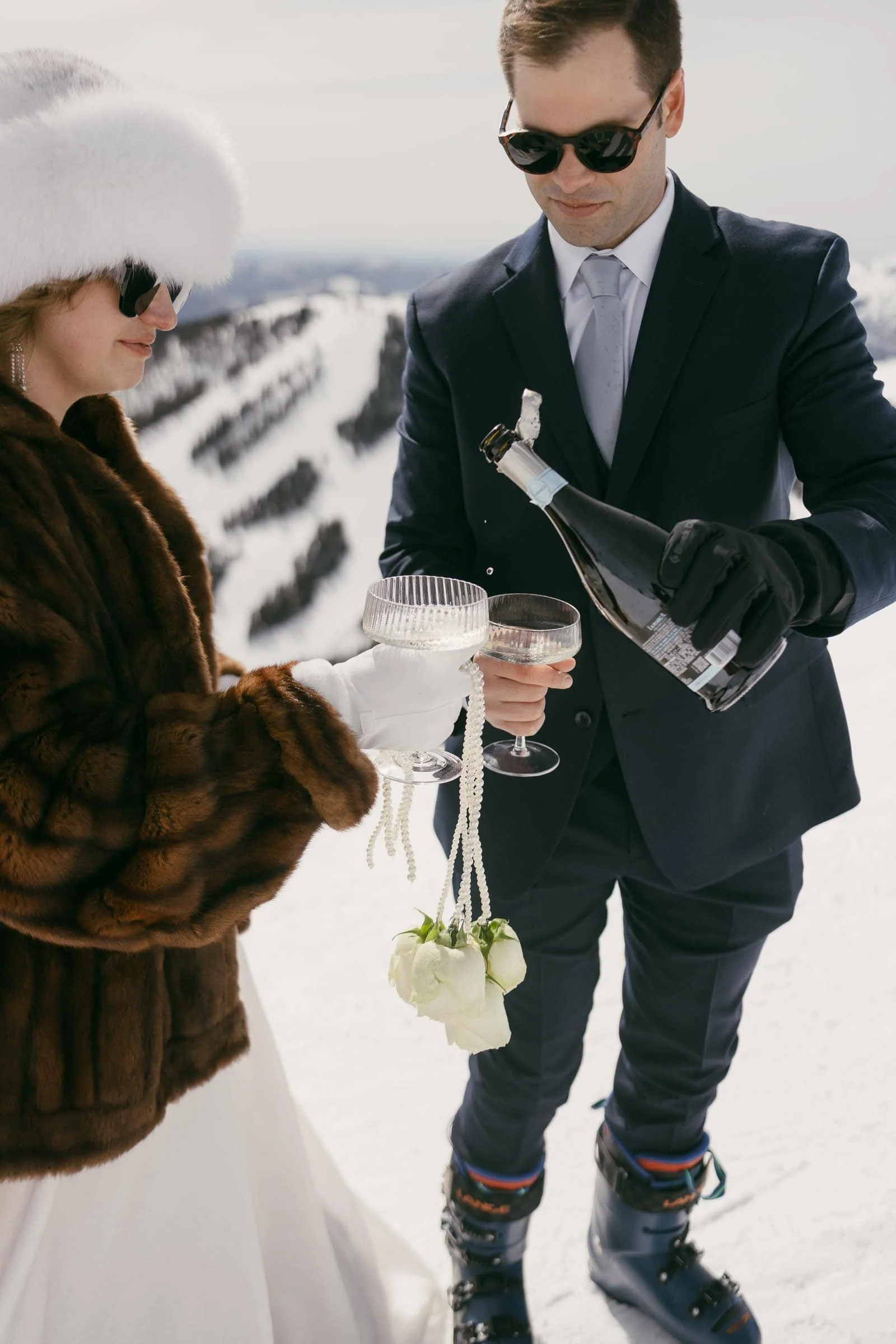  A couple dressed in formal winter attire, including sunglasses and gloves, stands on snowy mountains. The man pours champagne into two glasses, while the woman holds a small bouquet of white roses and a string of pearls. 