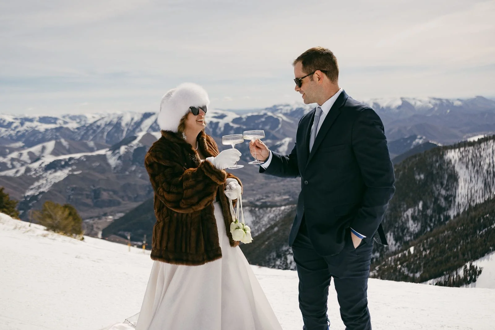  A bride in a fur coat and hat and a groom in a suit clink champagne glasses on a snowy mountain with scenic views and distant snow-covered peaks under a partly cloudy sky. 