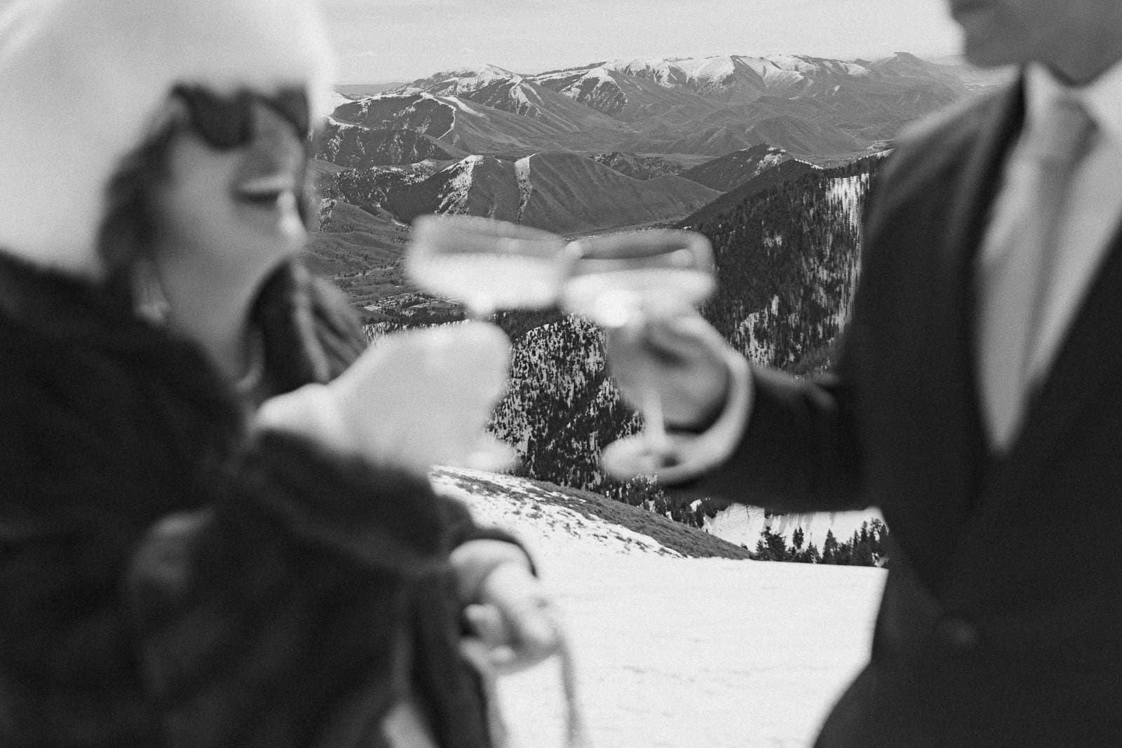 Two people dressed in winter attire raise champagne glasses together on a snowy mountain with scenic snow-covered peaks behind them. The black-and-white image is intentionally out of focus, creating a dreamy and artistic effect.