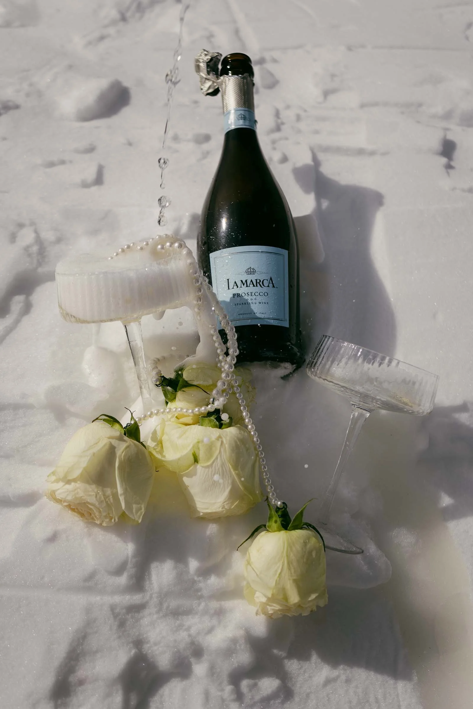  A bottle of La Marca Prosecco, two crystal champagne glasses, a strand of pearls, and three white roses are carefully arranged on the snow beneath bright sunlight, creating an elegant winter celebration scene. 