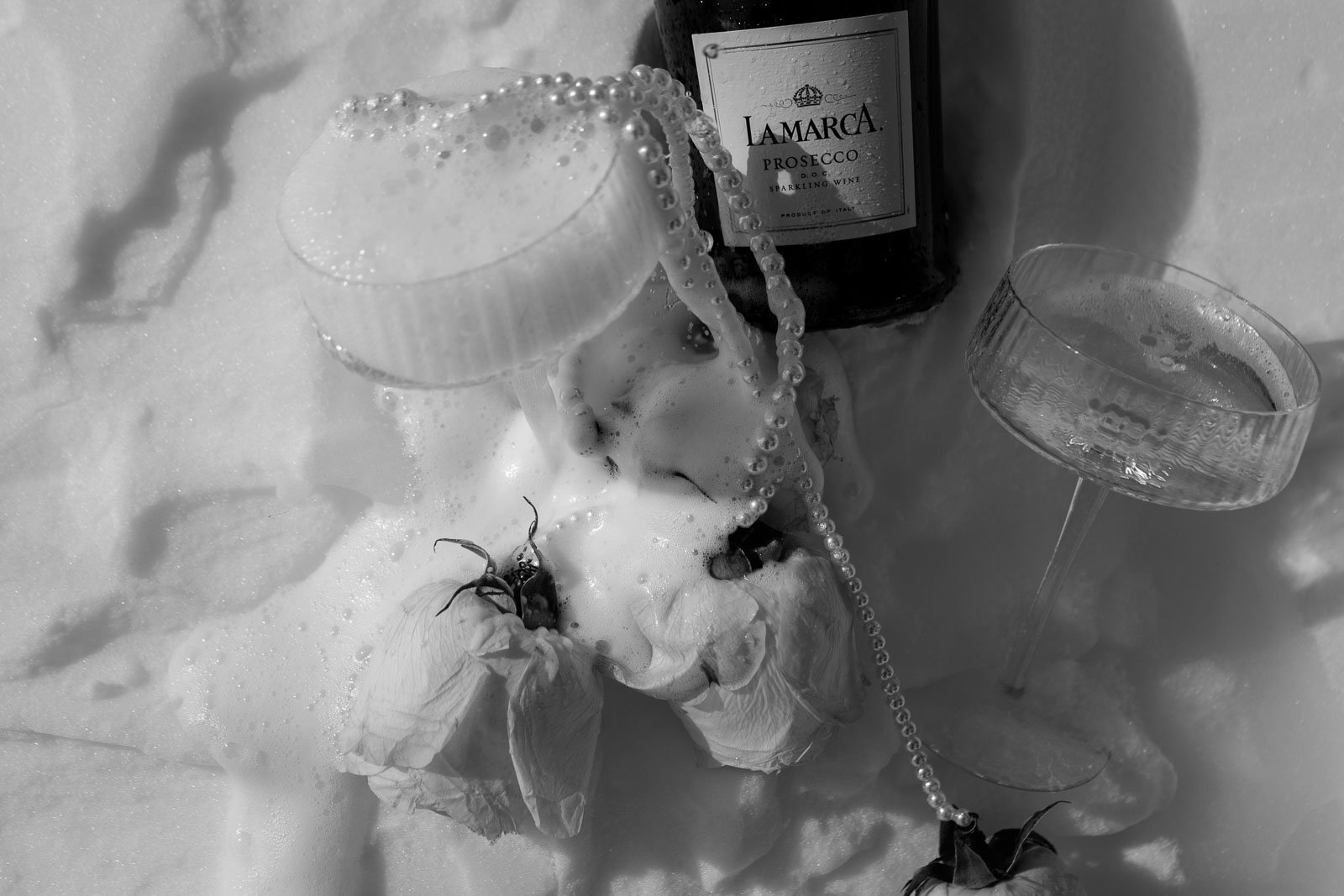  A bottle of La Marca Prosecco, two coupe glasses with bubbles, wilted roses, and a string of pearls are arranged on snow in black and white. 