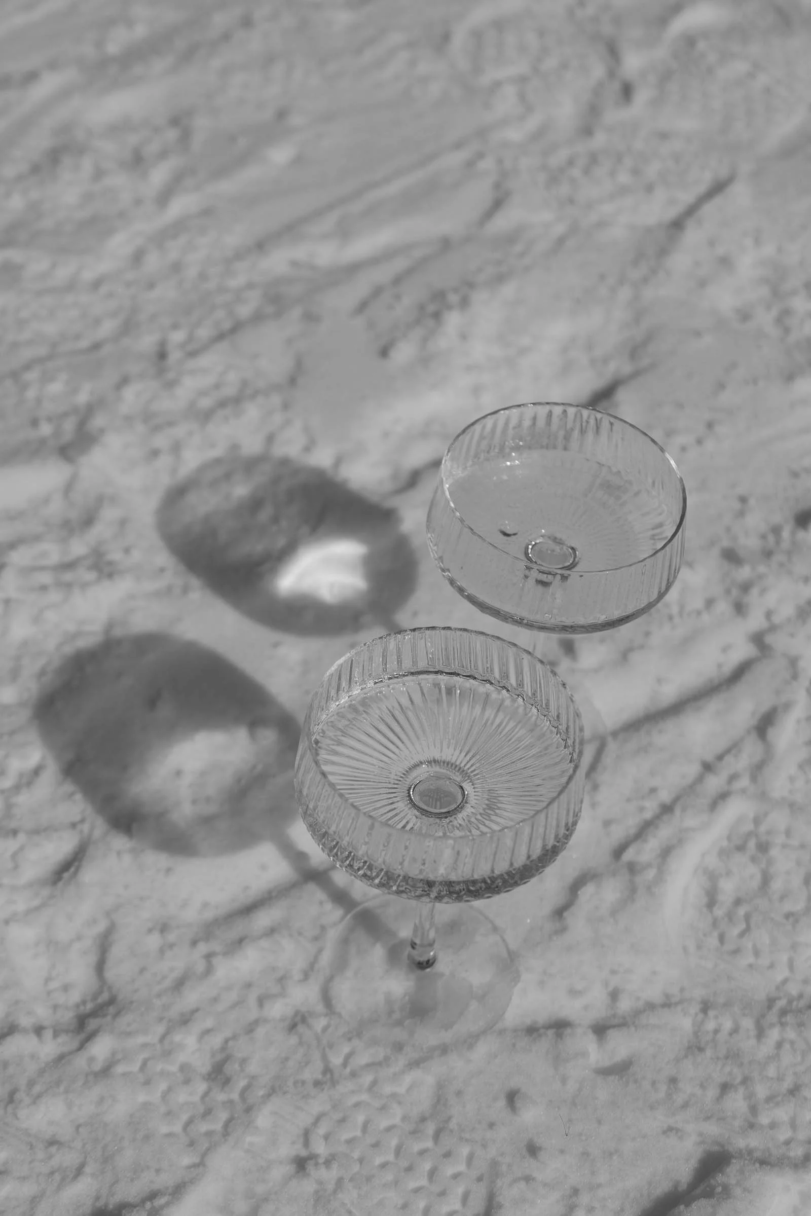  Two empty, textured coupe glasses casting distinct shadows on a sandy, patterned surface in black and white. 