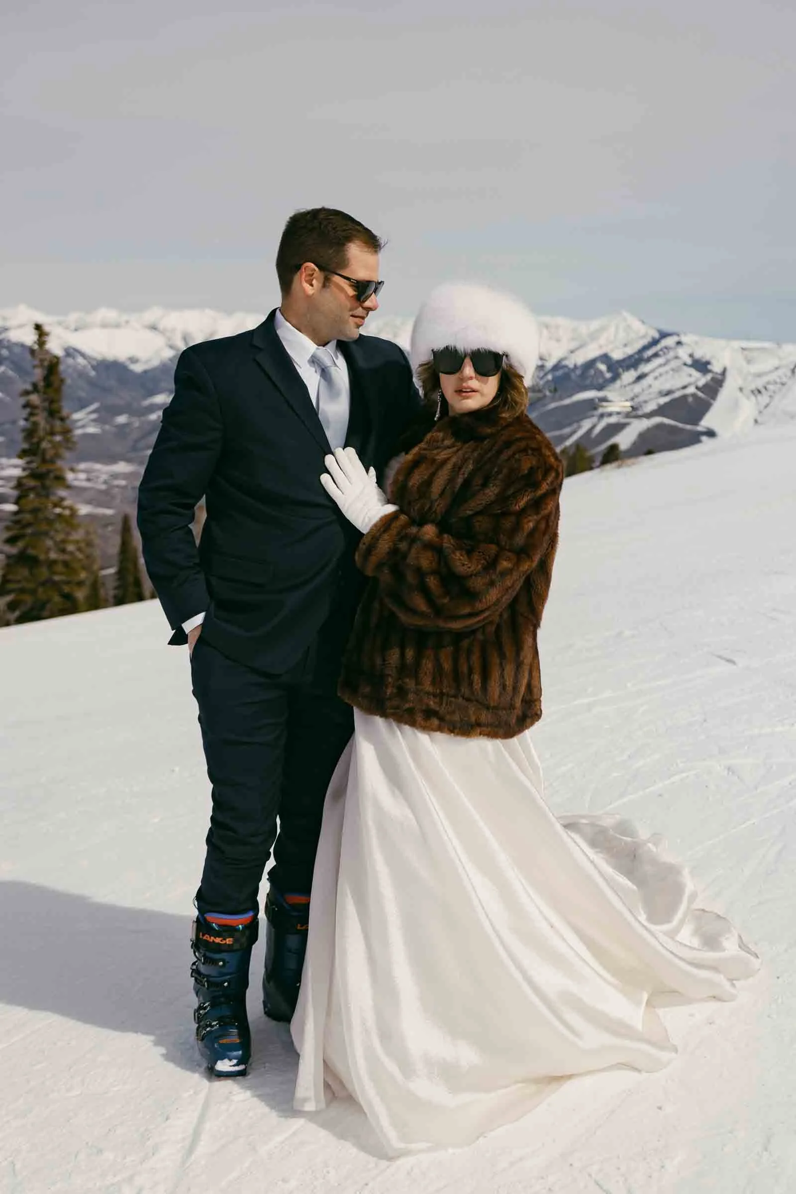  A bride in a white dress, fur coat, and hat stands on a snowy mountain next to a groom in a suit and ski boots. Both wear sunglasses; snow-covered peaks and trees are visible in the background. 