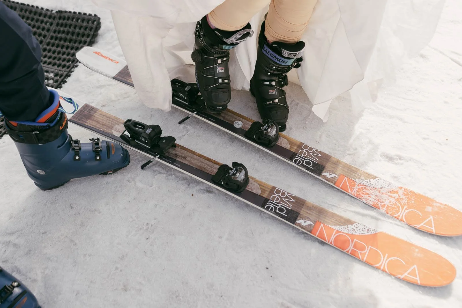  Close-up of two people in ski boots standing on skis in the snow; one person wears black boots and beige pants, the other wears blue boots. The skis are marked "Nordica" and have a wooden design with orange tips. 