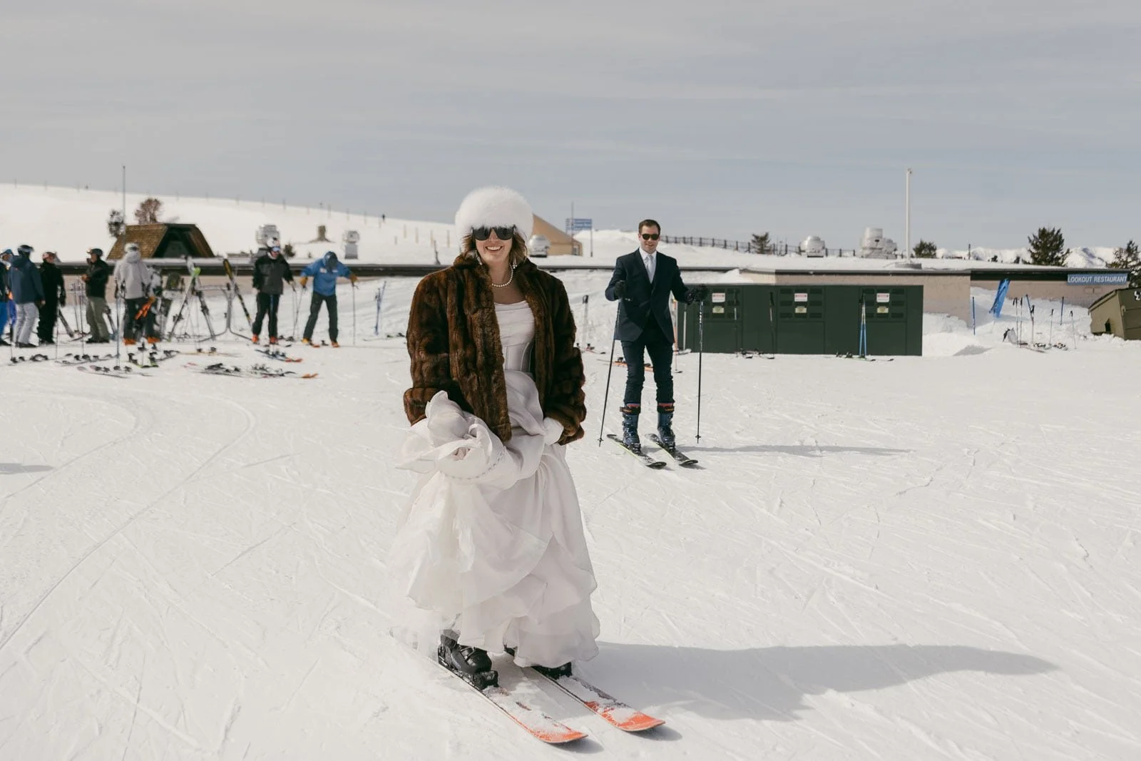 A person wearing a white dress, fur coat, and white winter hat skis across the snow while smiling. Another person dressed in a dark suit stands on skis behind them, with several skiers visible farther back in the snowy background.