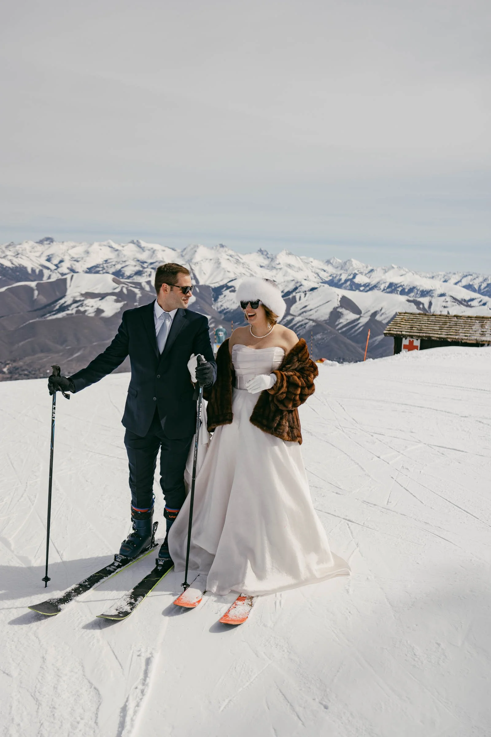  A bride in a white gown and fur shawl stands on skis next to a groom in a suit and sunglasses, both holding ski poles on a snowy mountain with snow-capped peaks in the background. 