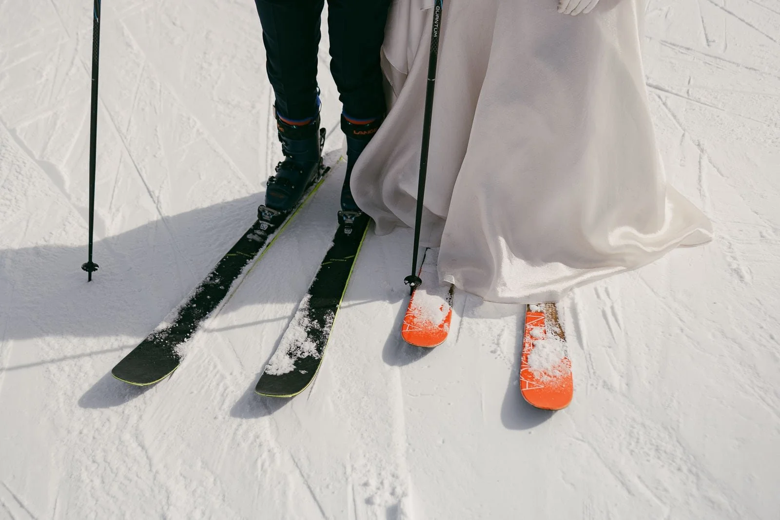  A couple stands together on the snow wearing ski gear; one person wears black pants with black skis, while the other wears a white dress paired with bright orange skis. Both hold ski poles as snow clings to parts of their skis and clothing. 
