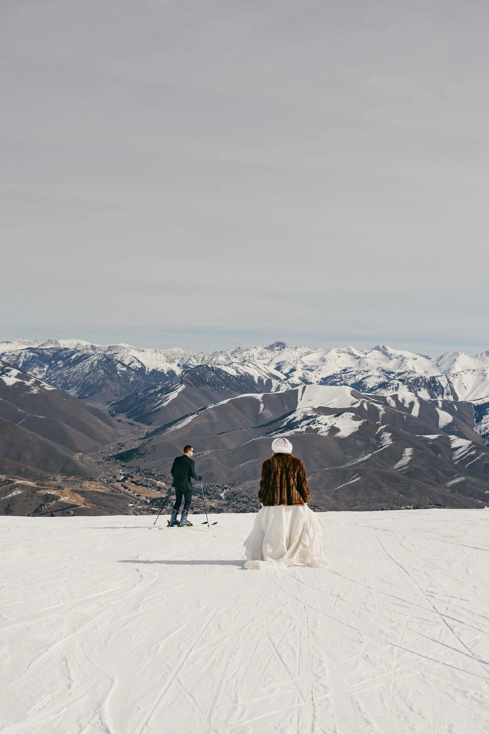  A person in a dress and fur coat stands on a snowy mountain, while another person on skis looks at the snow-capped mountain range under a cloudy sky. 