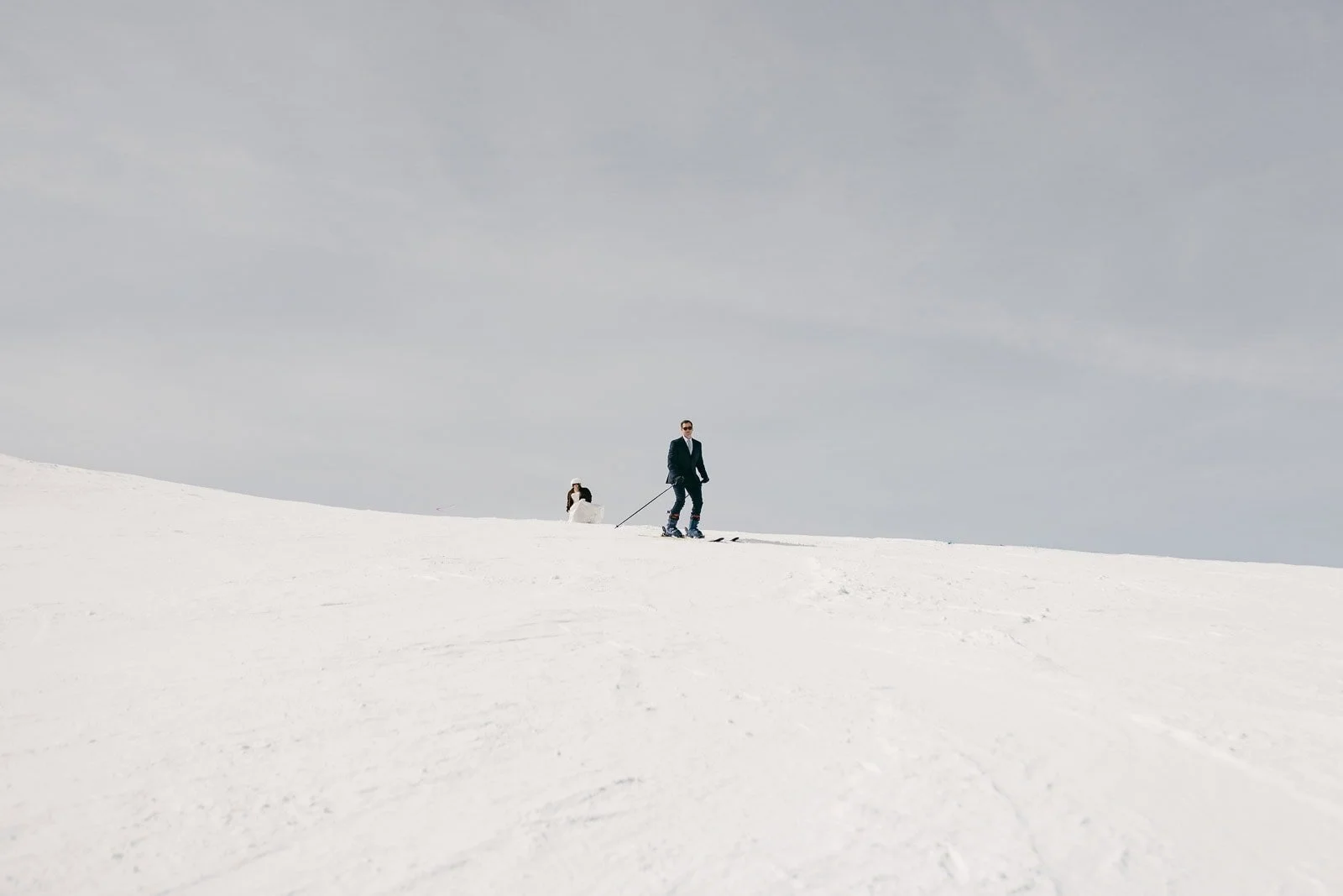  Two people ski across a snowy slope beneath a pale sky filled with light clouds. The minimal landscape feels expansive and open, with no trees or buildings visible anywhere in the scene. 