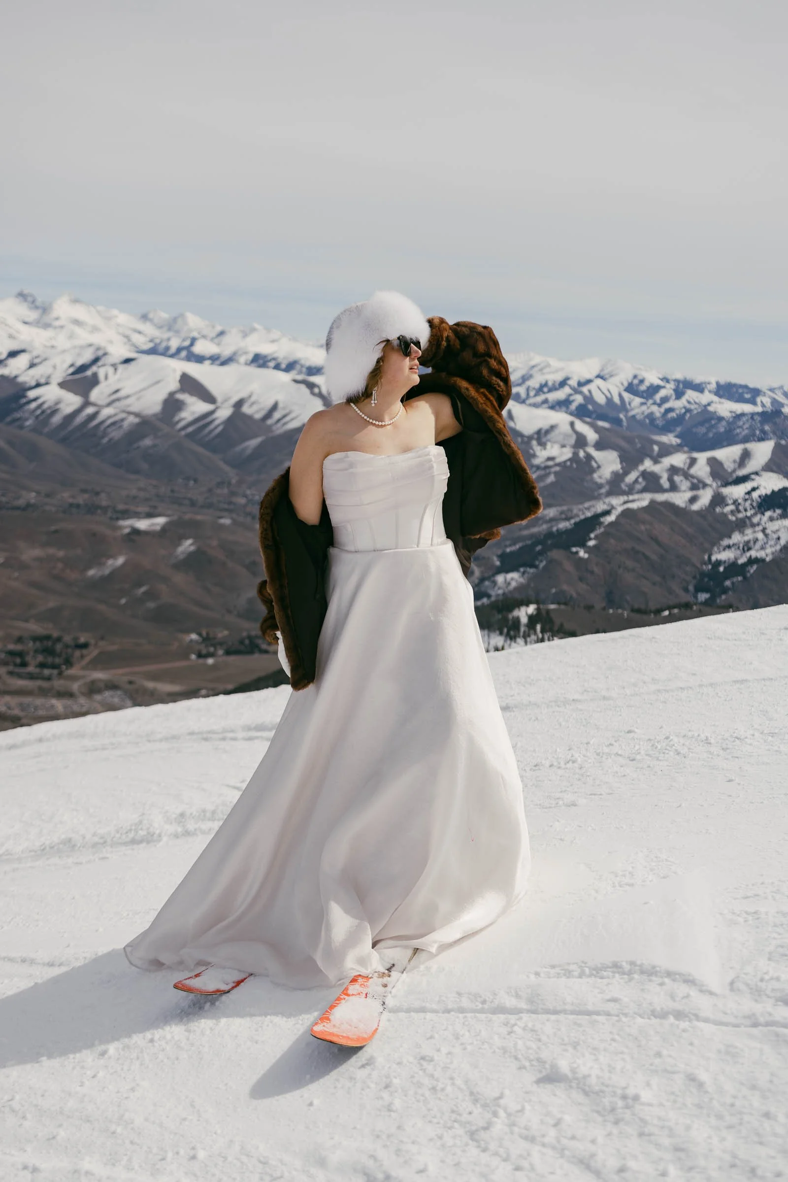  A person wearing a strapless white gown, fur hat, and brown coat stands on a snowy mountain slope, with snow-covered peaks and ridges in the background. They are also wearing pink snowshoes. 