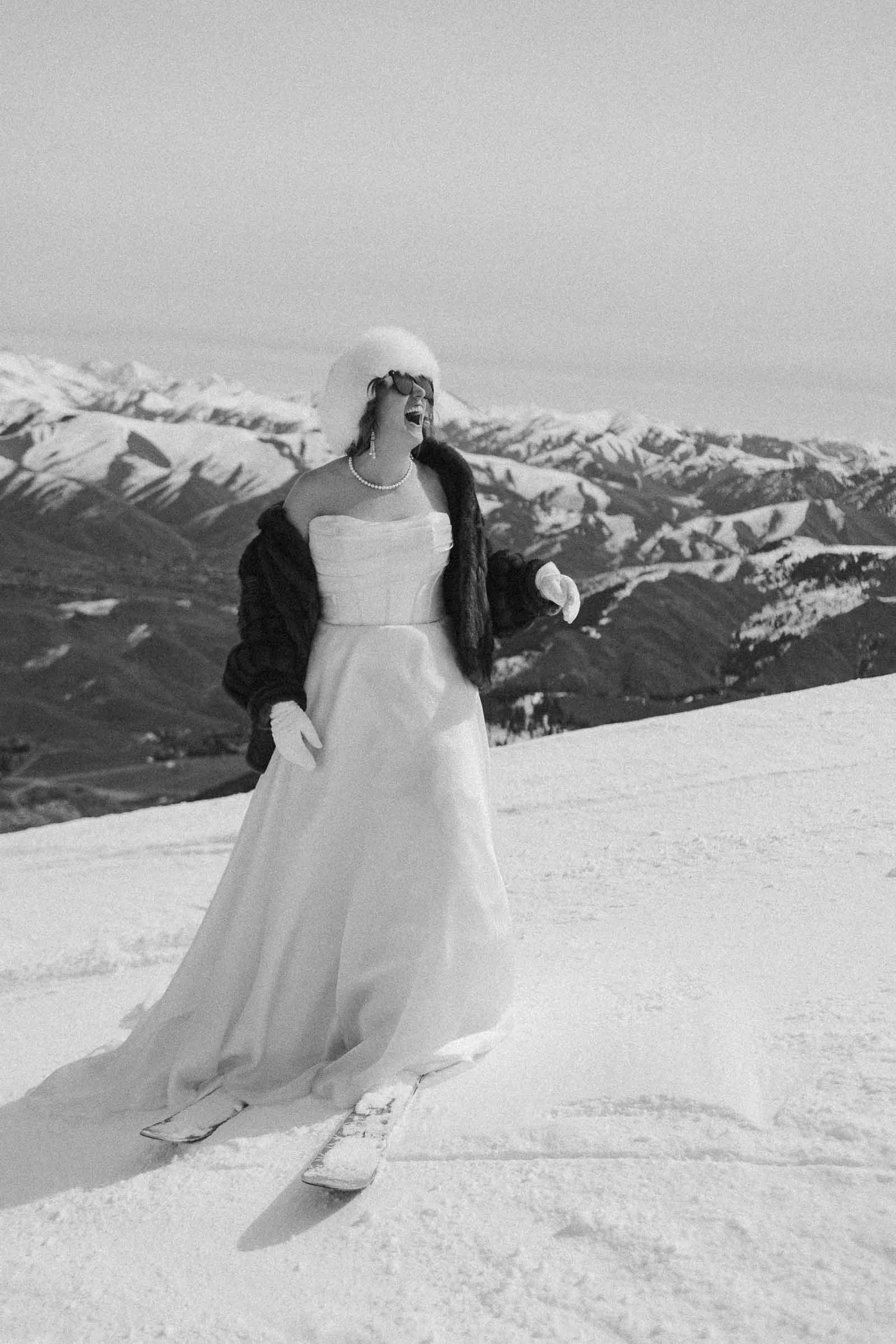  A person in a strapless white gown, fur hat, and coat stands on skis in the snow, surrounded by snowy mountains under a clear sky. The image is in black and white. 