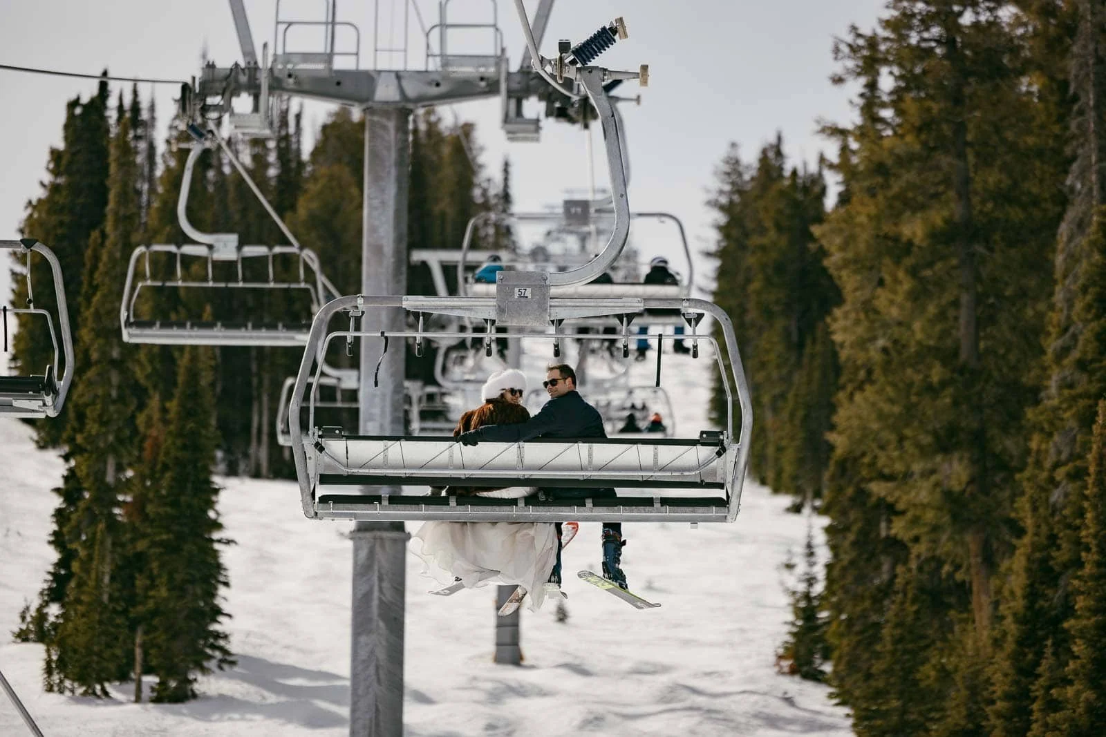 A couple rides together on a ski lift above snow-covered ground and tall pine trees, making their way up the mountain while other skiers can be seen in the snowy background below.