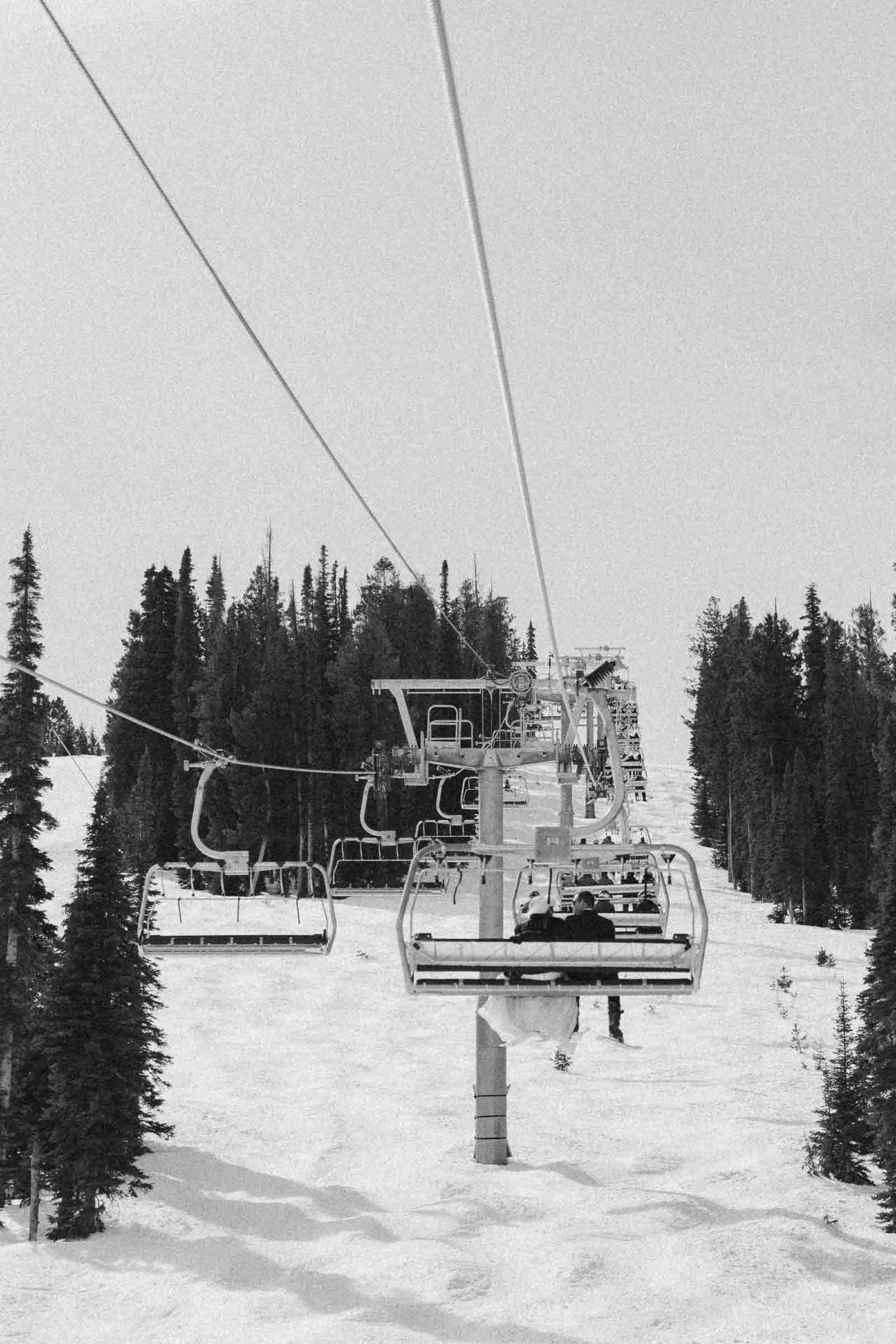  A black and white photo shows people riding a ski lift over a snowy slope, surrounded by tall pine trees on both sides under a clear sky. 